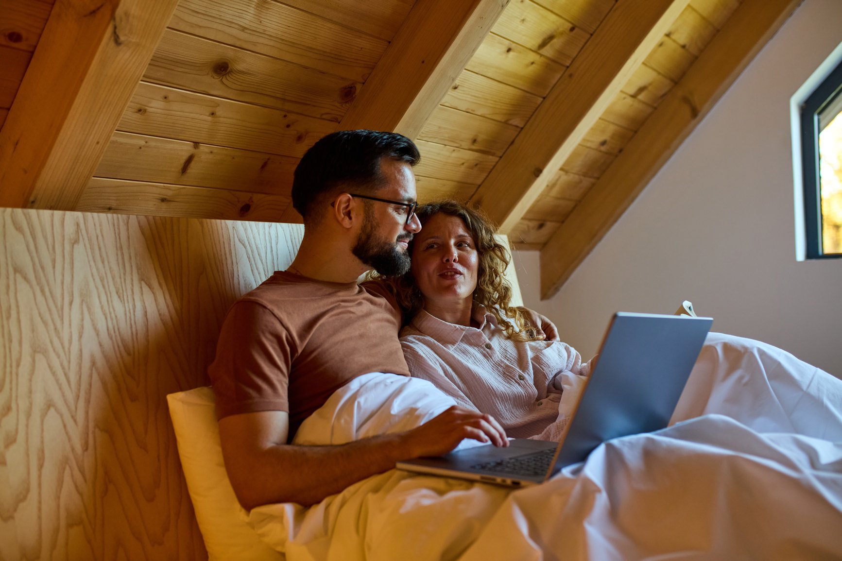 A couple sitting in bed in a cozy, wood-paneled bedroom, looking at a laptop together. EFT Couples Therapy in Royal Oak, Michigan and online statewide.