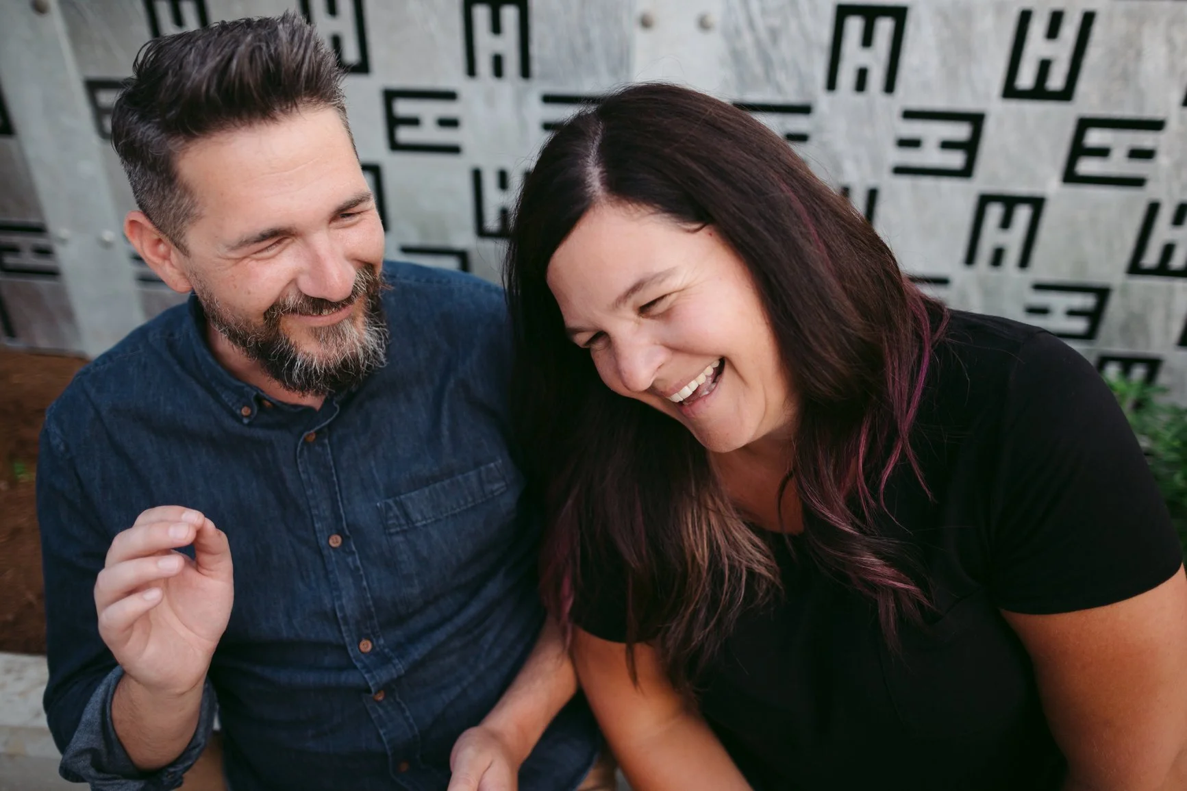 A man and a woman sharing a joyful moment, sitting close together and laughing with their heads touching, in a casual setting near a wall with a geometric black and gray pattern. EFT Couples Therapy in Royal Oak, Michigan and online statewide