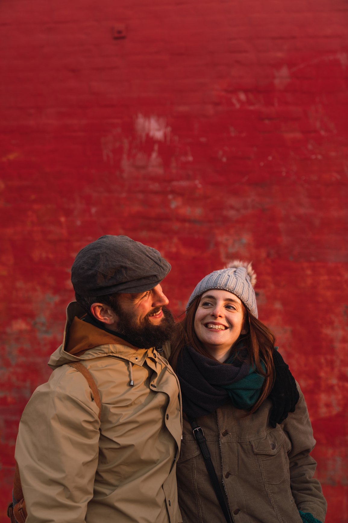 A man and woman standing close together, smiling. The man has a beard, wearing a gray cap and tan jacket. The woman has long hair, wearing a gray beanie with a pom-pom, a green scarf, and a brown jacket. EFT Couples Therapy in Royal Oak, Michigan