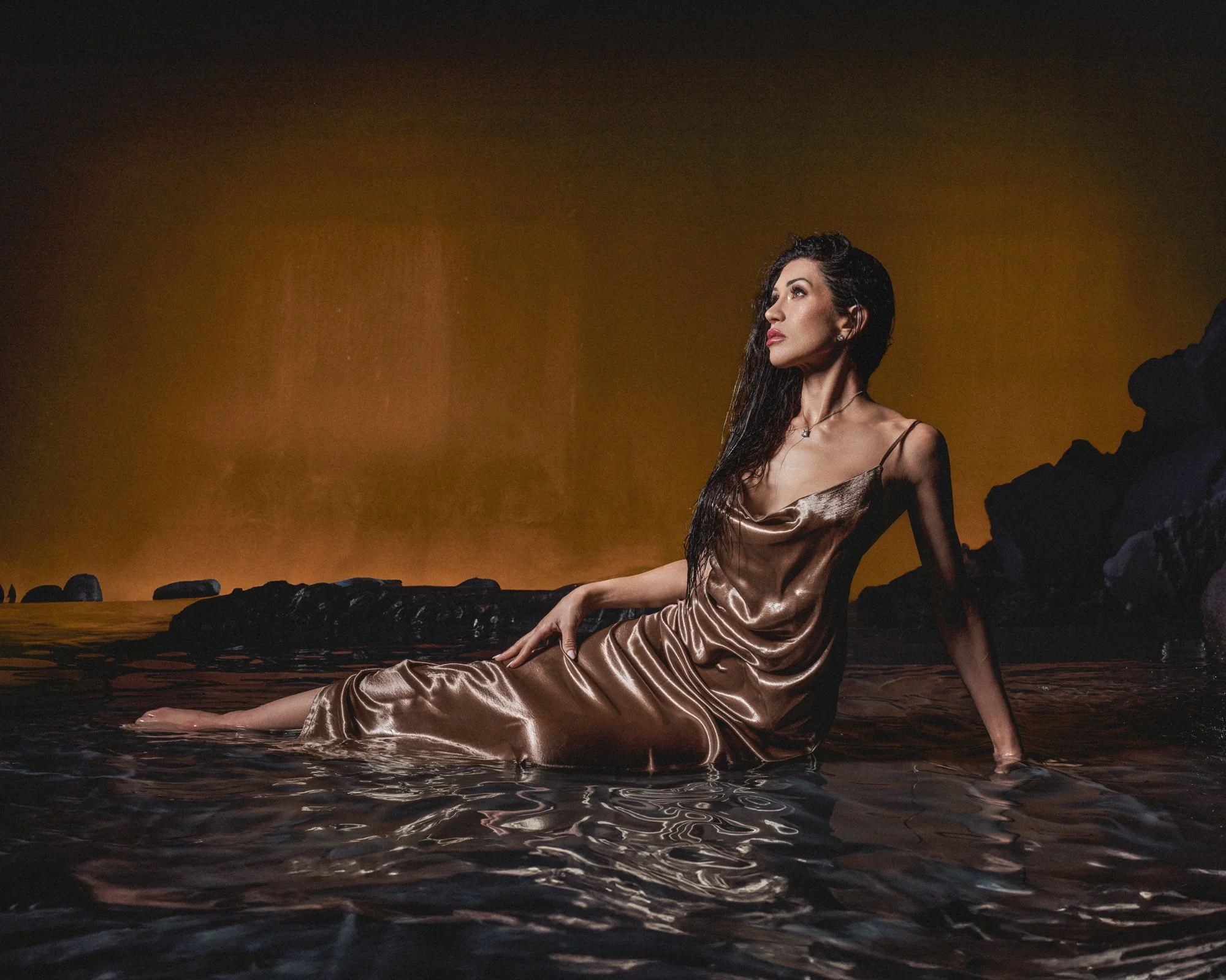 A woman with long dark hair in a satin dress sitting in shallow water near dark rocks during sunset with an orange sky.