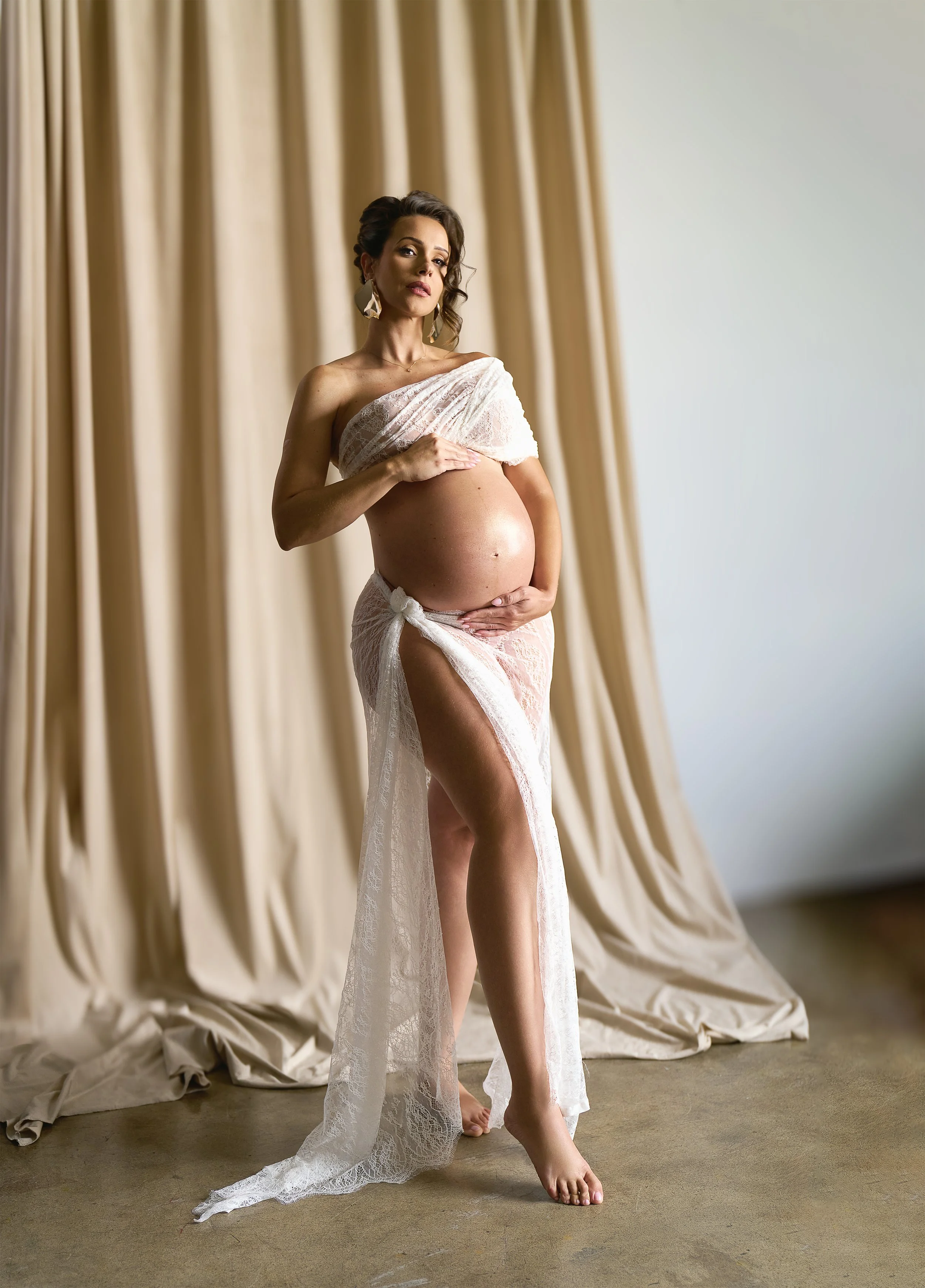 a pregnant woman wearing fine lace, standing effortlessly with a foraml up-do basking in sun while softly looking at the camera in ahigh fashion style