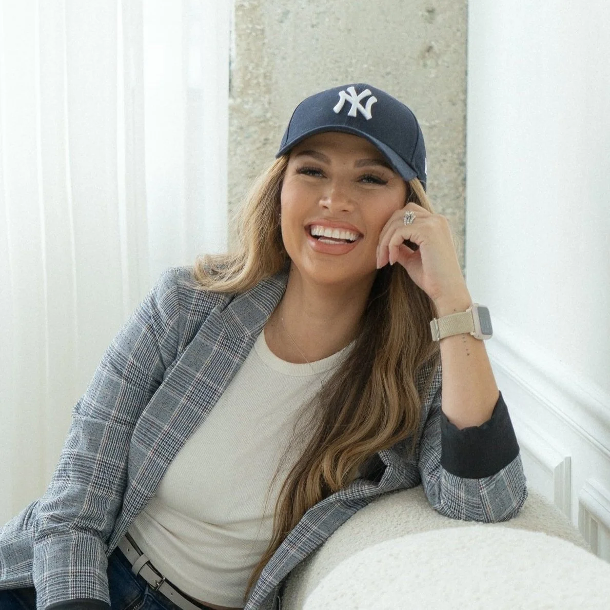 A smiling woman wearing a navy New York Yankees baseball cap, plaid blazer, white t-shirt, and smartwatch, sitting in a bright, modern indoor setting.