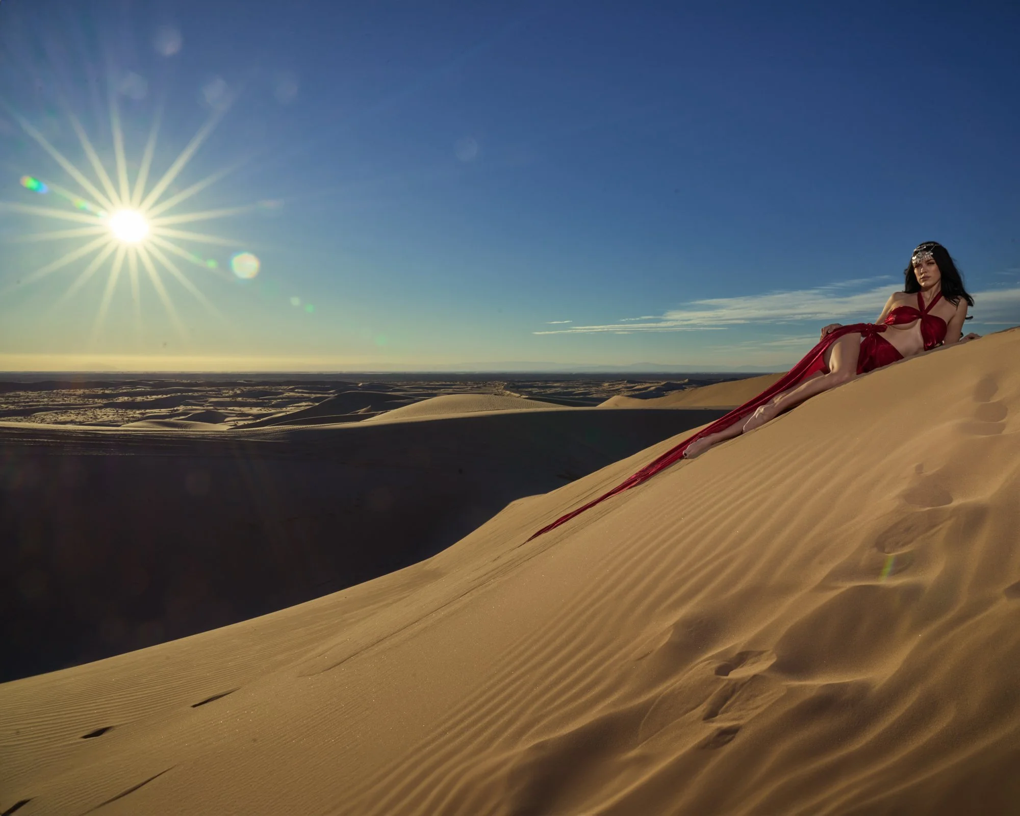 A woman in a red dress lying on a sand dune in a desert landscape with the sun shining brightly in the sky.