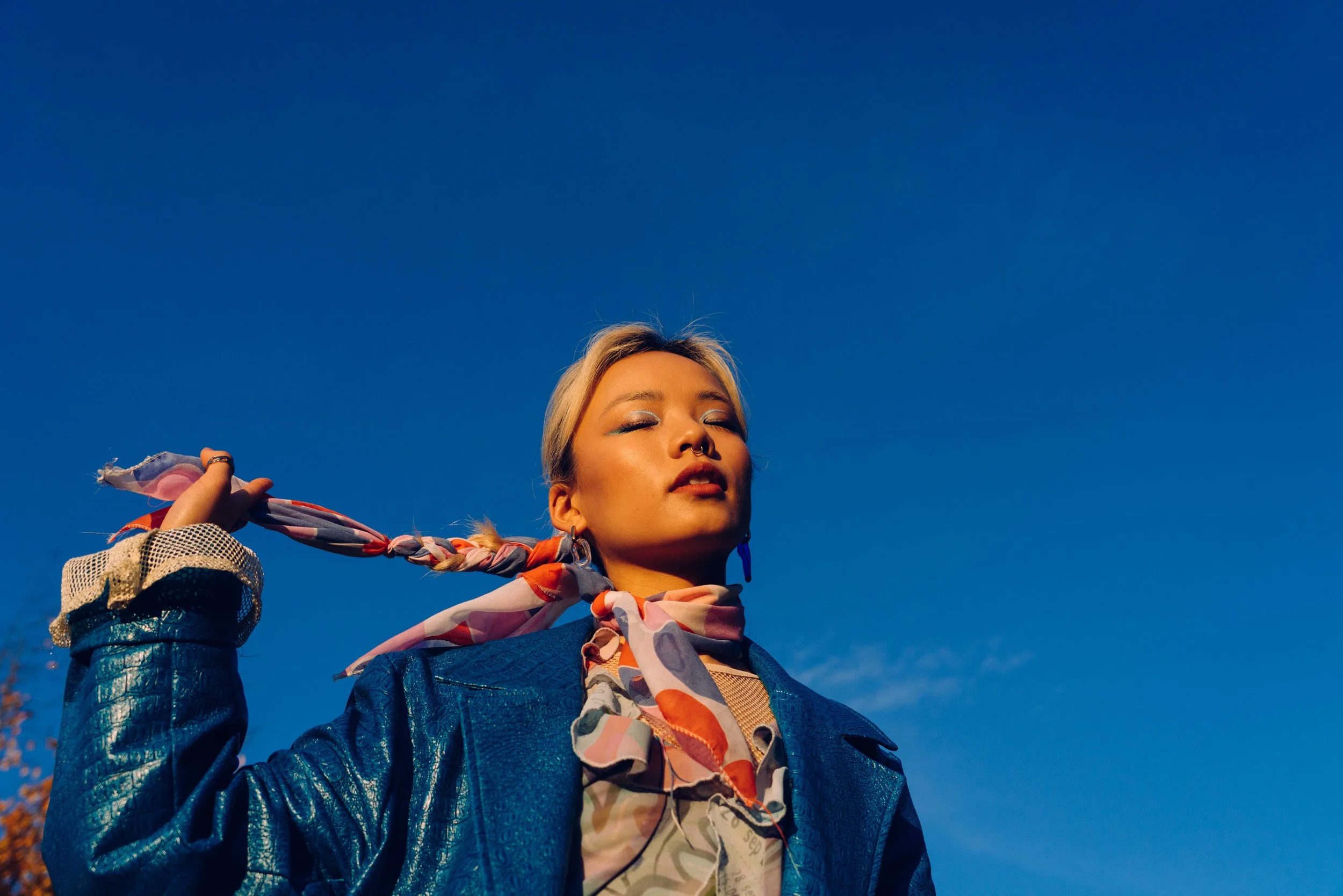Junge Frau mit bunter Halsschleife, blauer Jacke, Sonnenbrille und offener Augen, die die Augen schließt, unter klarem blauen Himmel.