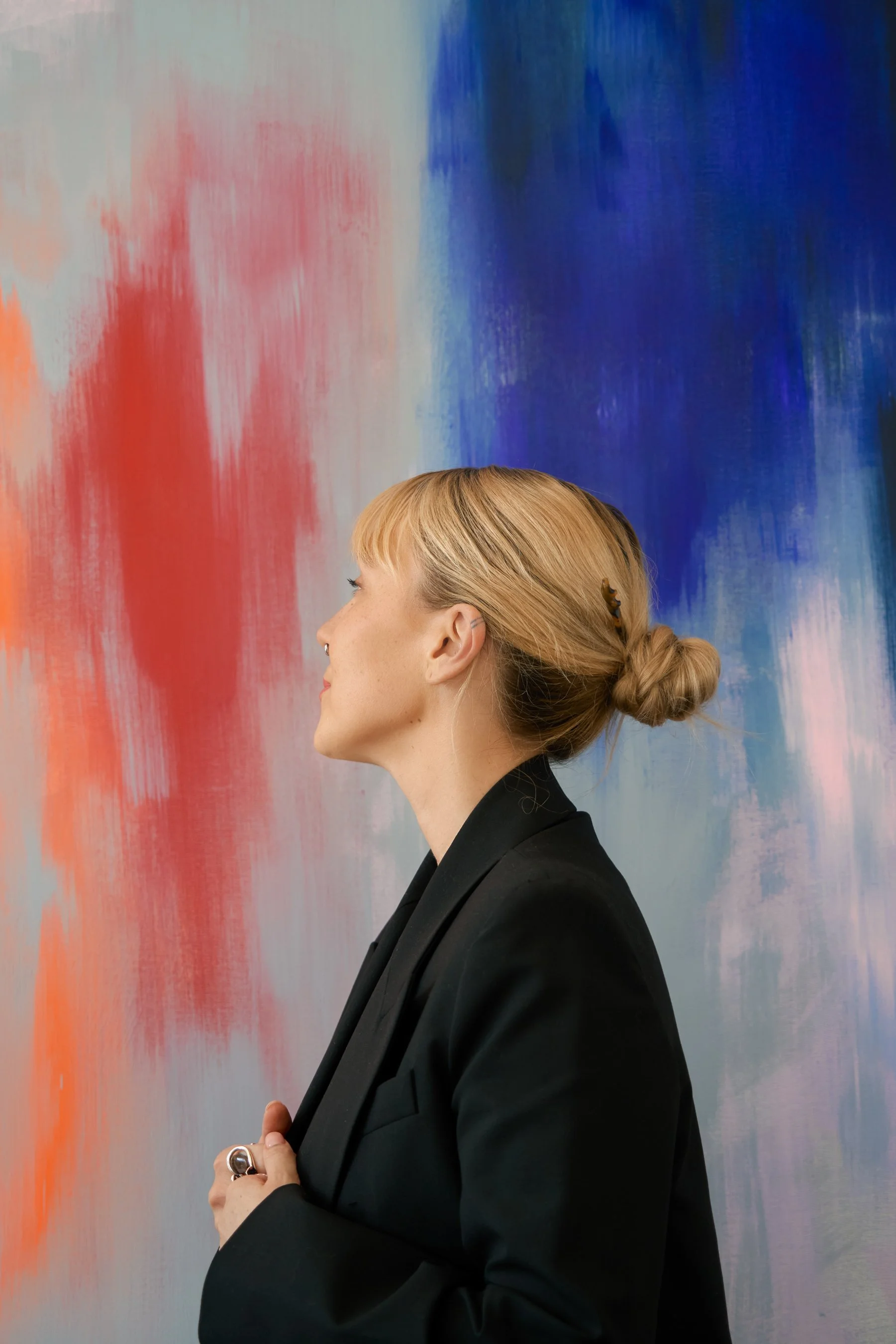 Seitliche Profile eines blonde Frau in schwarzem Blazer vor einem abstrakten Gemälde mit blauer, roter und weißer Farbgestaltung.