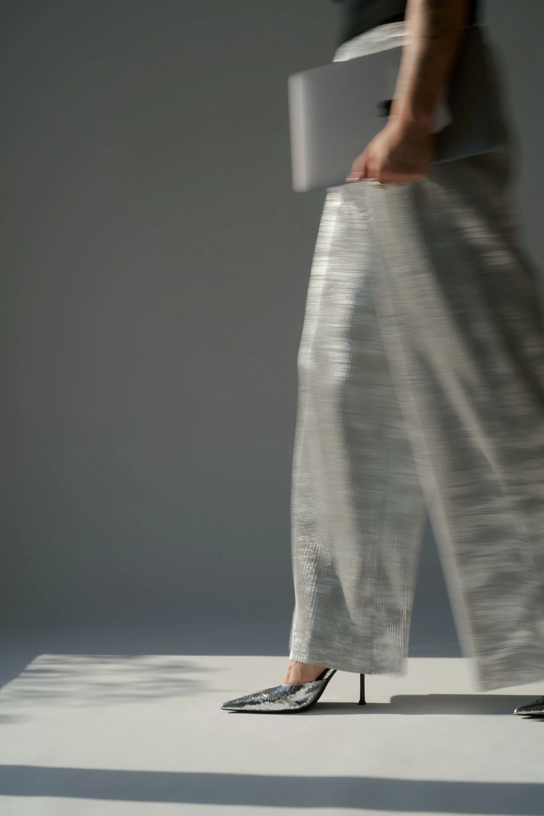 Eine Person trägt elegante, glänzende silberne Schuhe mit hohen Absätzen und eine schimmernde, gemusterte Hose. Die Person hält ein Tablet in der Hand. Der Hintergrund ist grau.