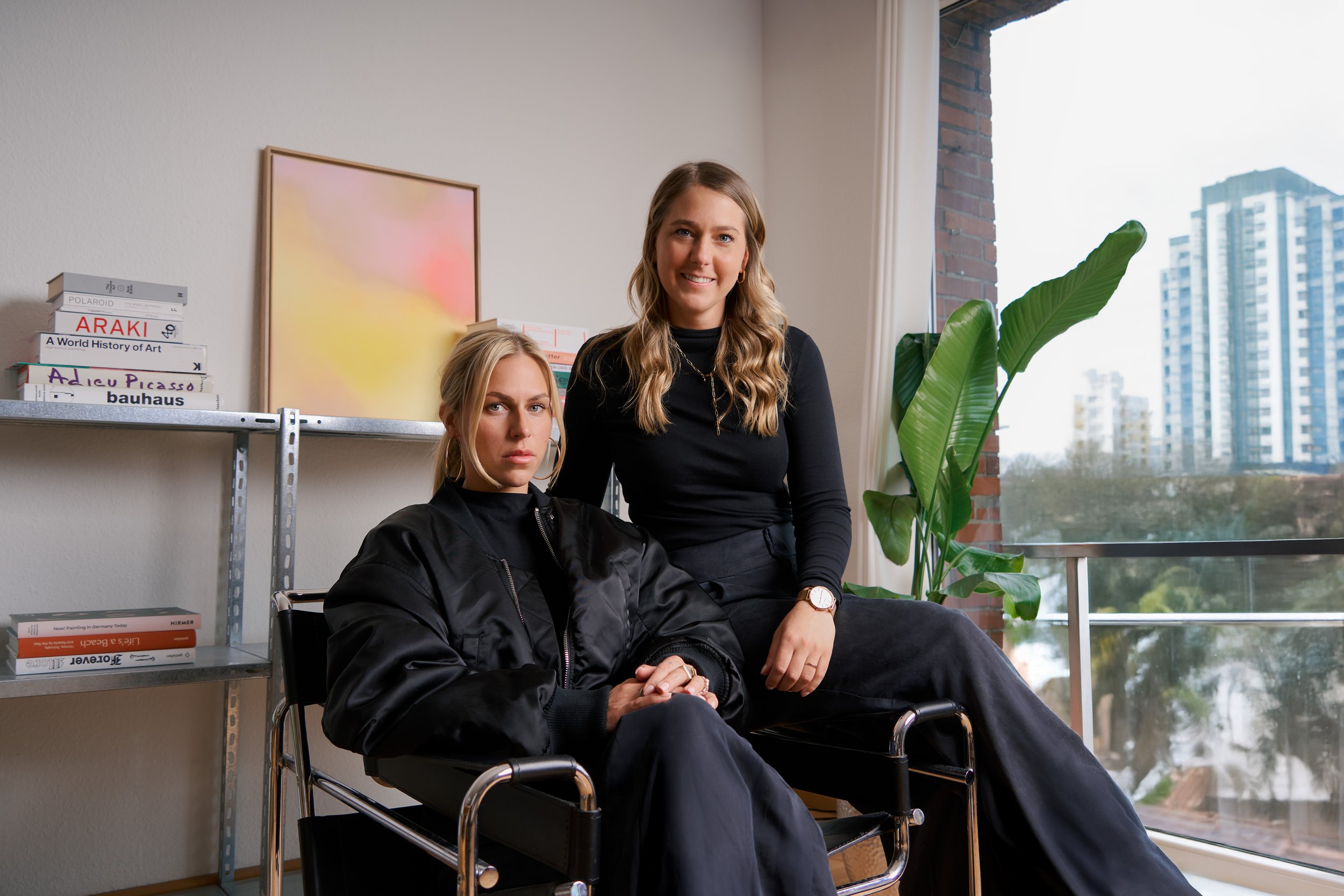 Zwei junge Frauen mit blondem Haar, in einem modernen Raum, sitzend und stehend vor einer weißen Wand mit Büchern und Kunstwerken, neben einem großen Fenster mit Blick auf Hochhäuser, einer Pflanze und einer Stehlampe.