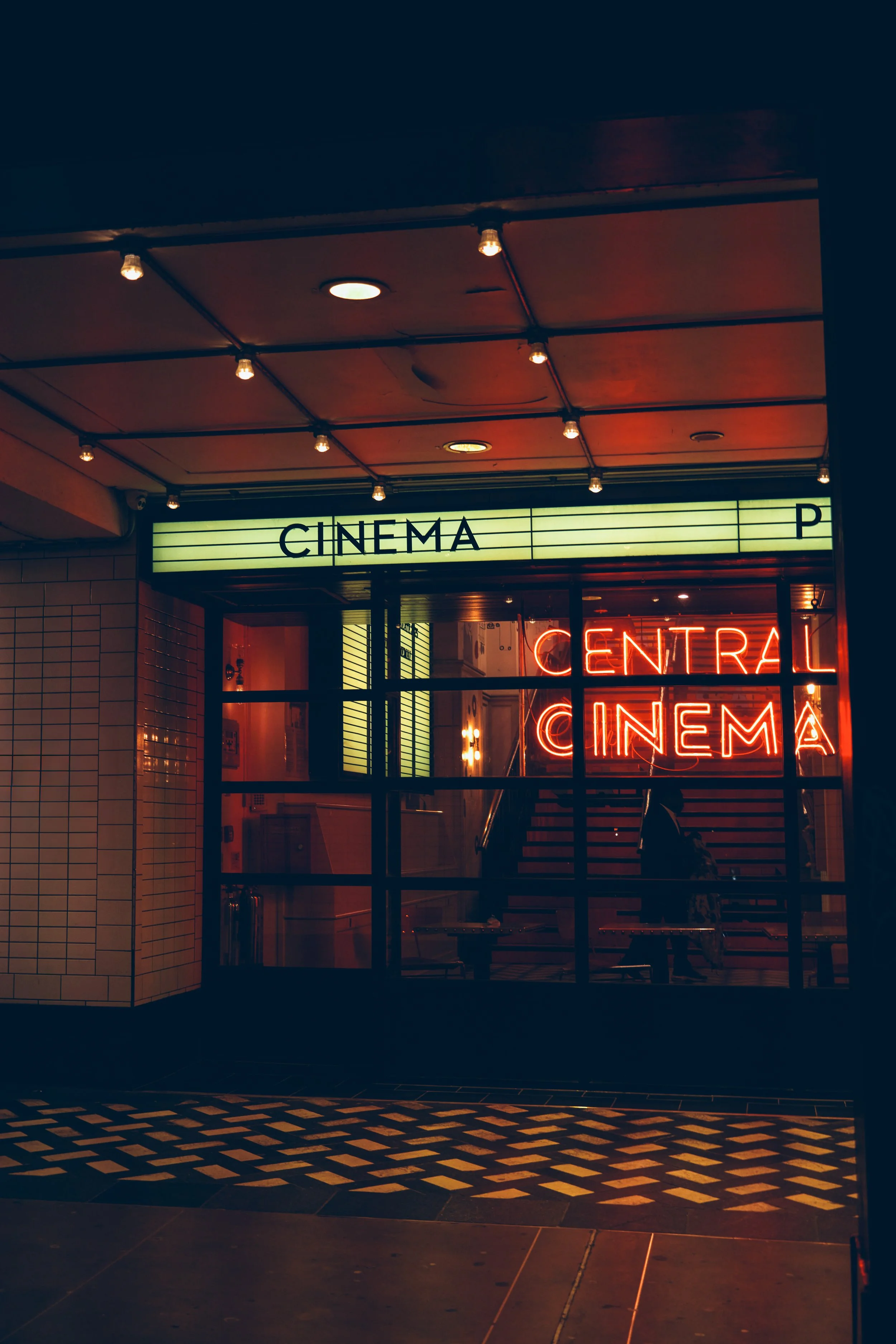 Außenansicht eines Kinos bei Nacht mit beleuchteten Neon-Schildern, die 'CINEMA' und 'CENTRAL' anzeigen. Das Kino hat große Fenster, durch die Sitzbereiche sichtbar sind, und eine beleuchtete Decke mit mehreren Glühbirnen.