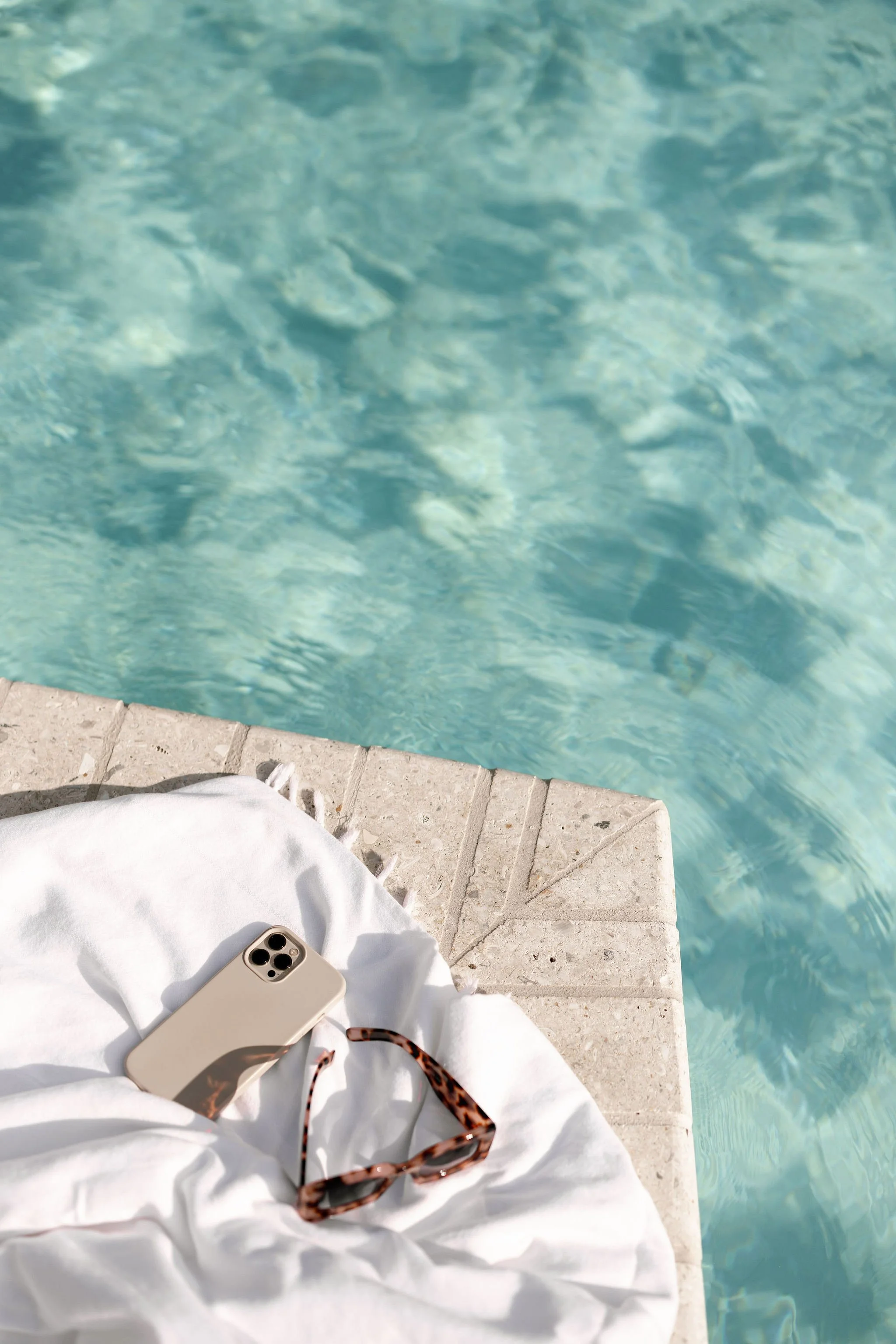 Nahaufnahme eines Handys, einer Sonnenbrille und eines weißen Kleidungsstücks auf einer Steintreppe neben einem Pool.