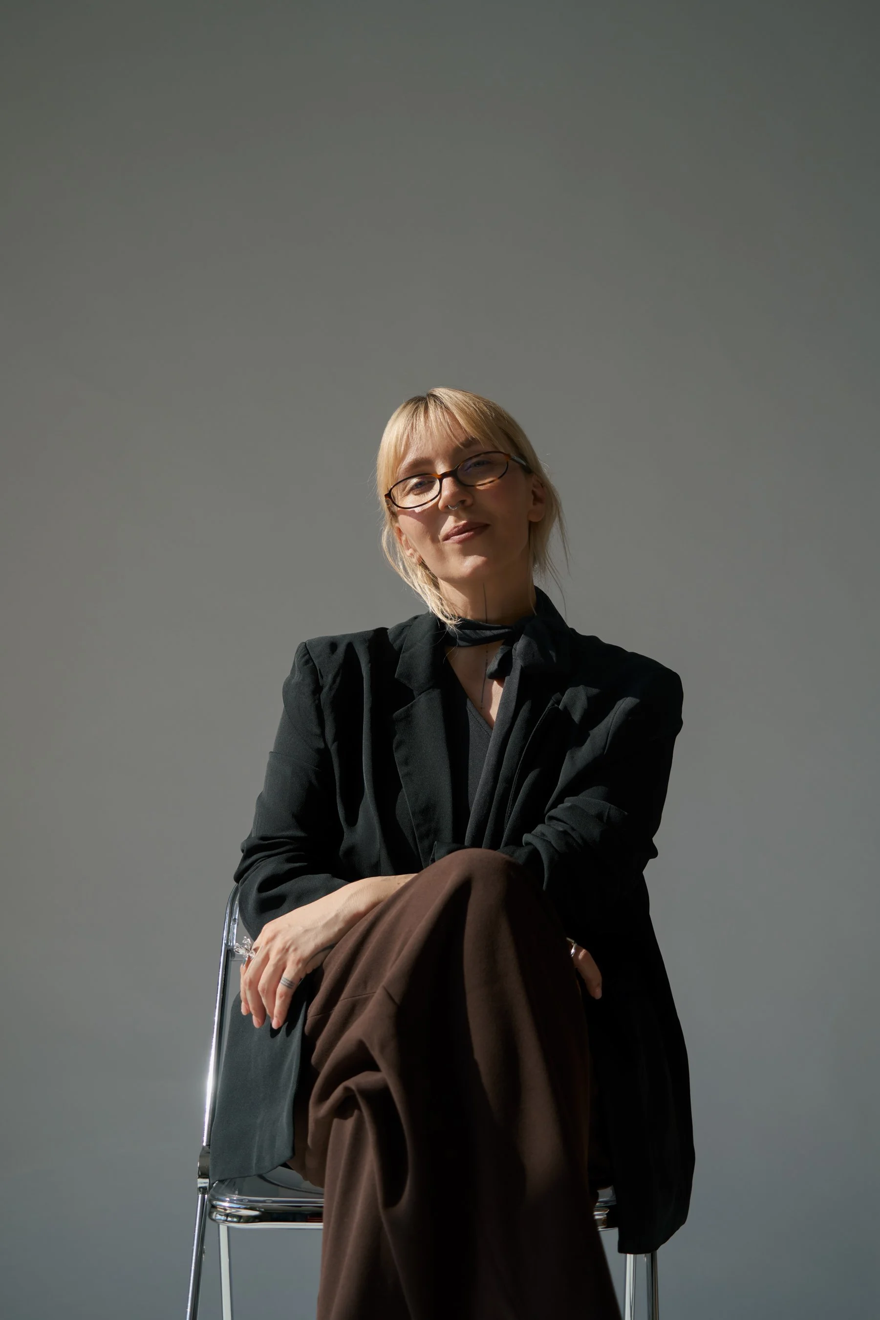 Eine Frau mit blonden Haaren, Brille und dunkler Kleidung sitzt auf einem Stuhl vor einem grauen Hintergrund. Sie schaut leicht lächelnd in die Kamera.