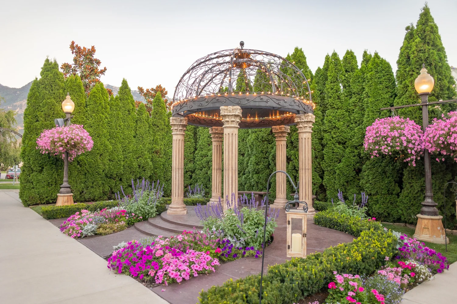 The garden courtyard at Le Jardin — a stone gazebo lit with warm lights, surrounded by manicured topiaries and beds of pink and purple flowers, with the Wasatch mountains beyond