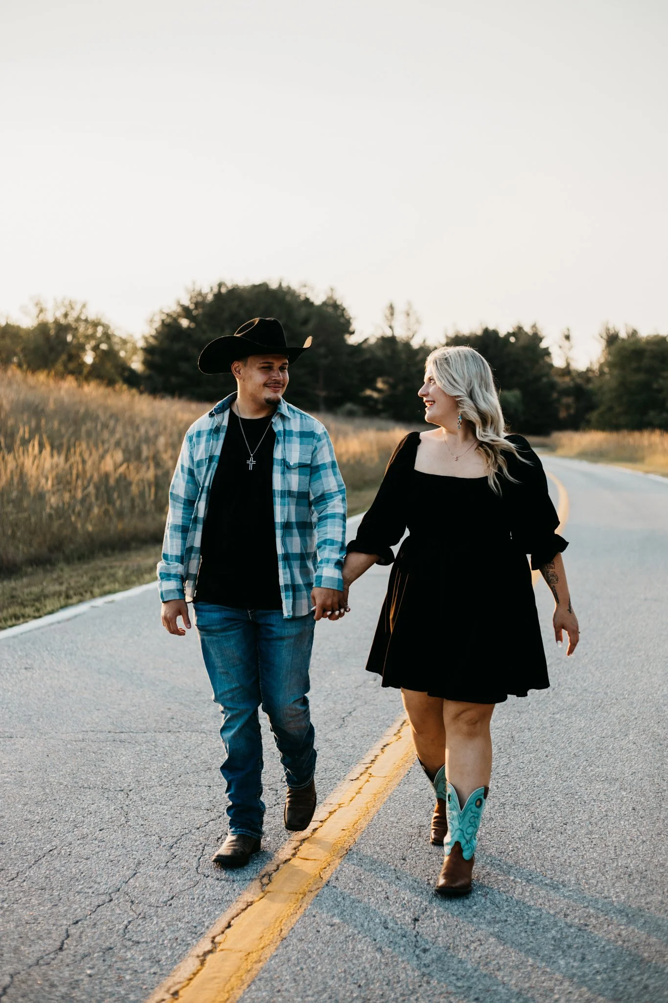 A couple walking hand in hand down a rural road during sunset; the man wears a cowboy hat, plaid shirt, and jeans, while the woman wears a black dress with cowboy boots and turquoise accents, smiling at each other.