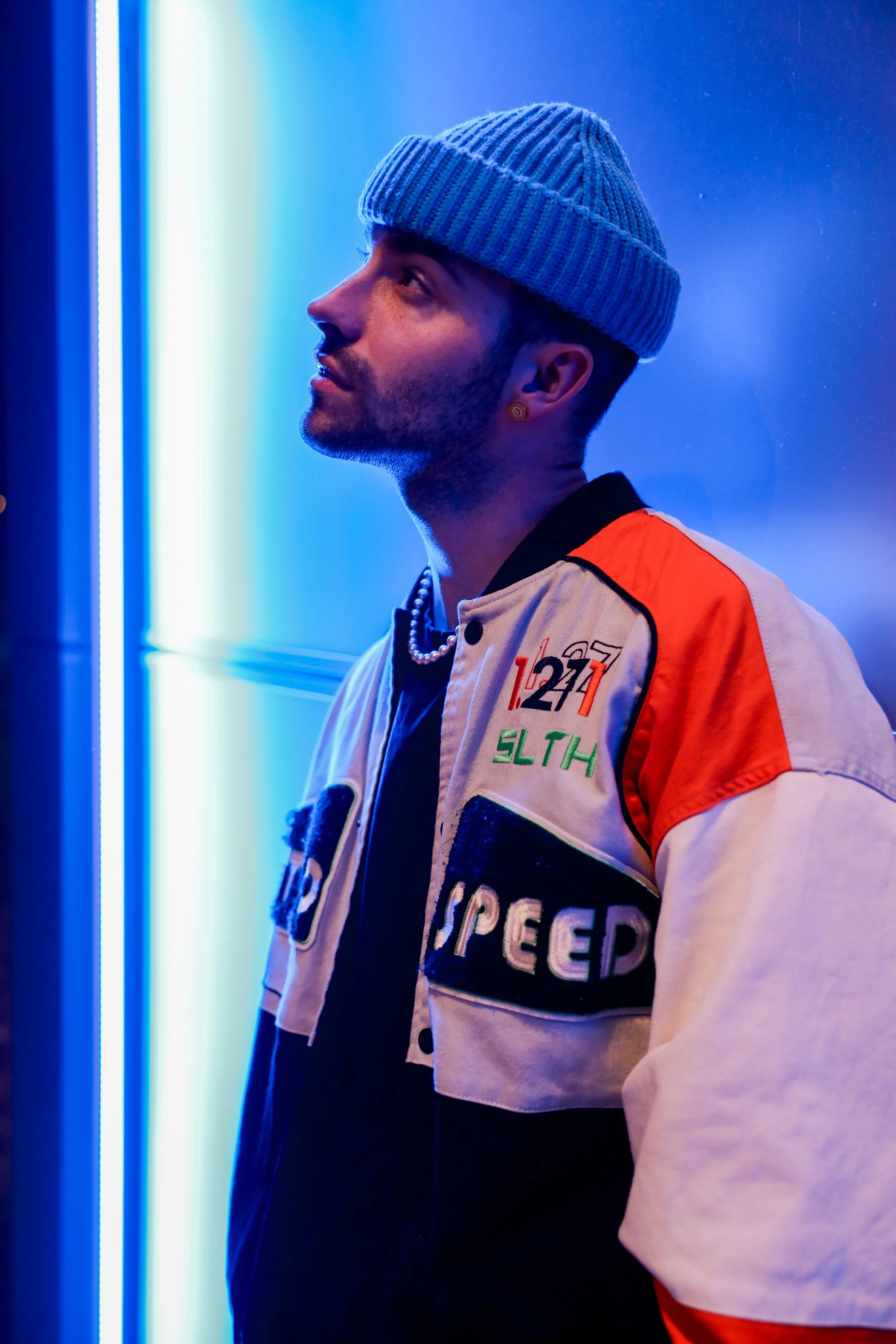 A young man with a beard and mustache wearing a gray knit beanie, a pearl necklace, and a beige and orange jacket with patches and text, standing in front of a blue-lit background, looking to his left.