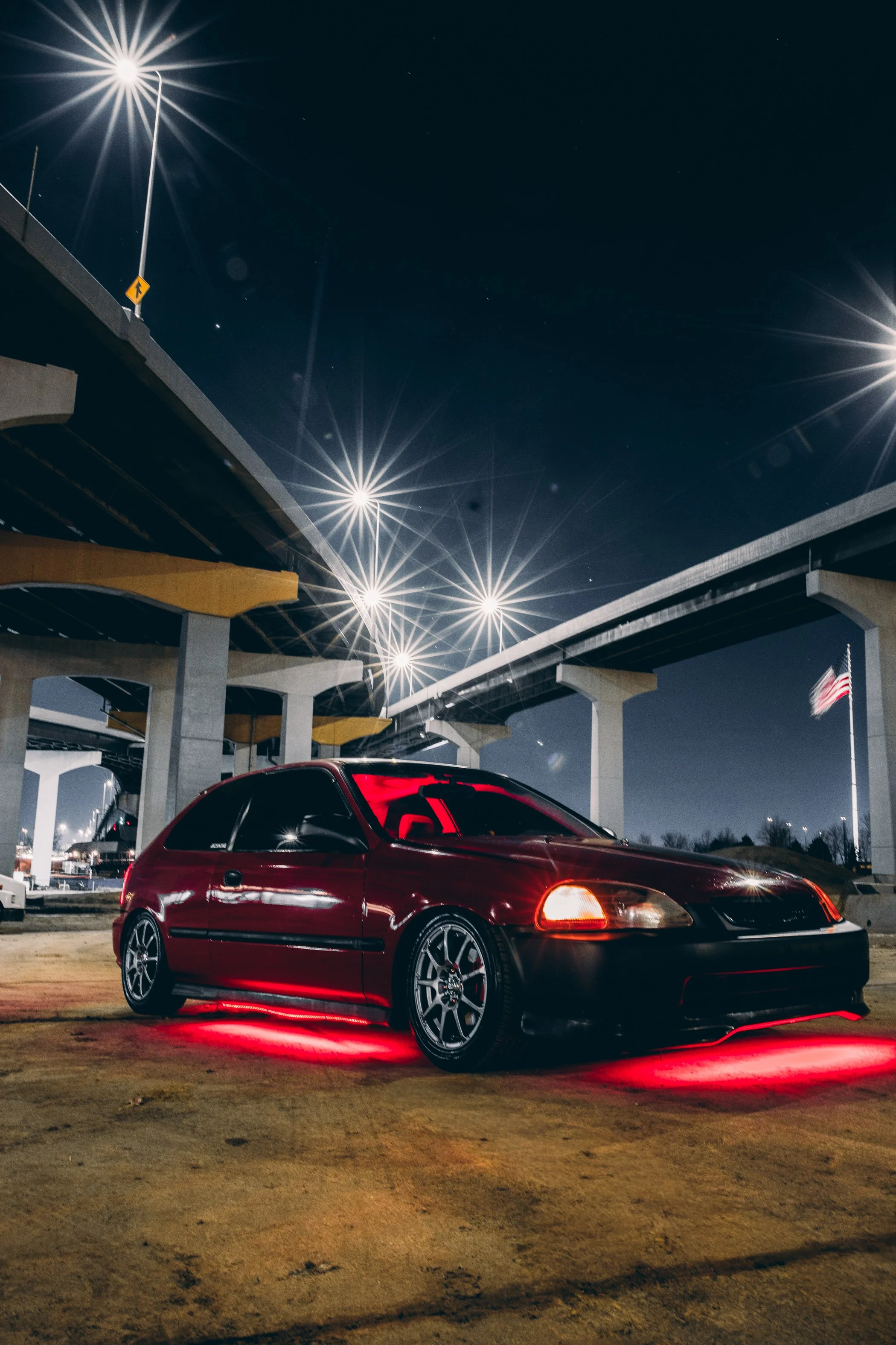 A red sports car with custom wheels and red underglow lights parked under a bridge at night, with bright streetlights and a dark sky in the background.