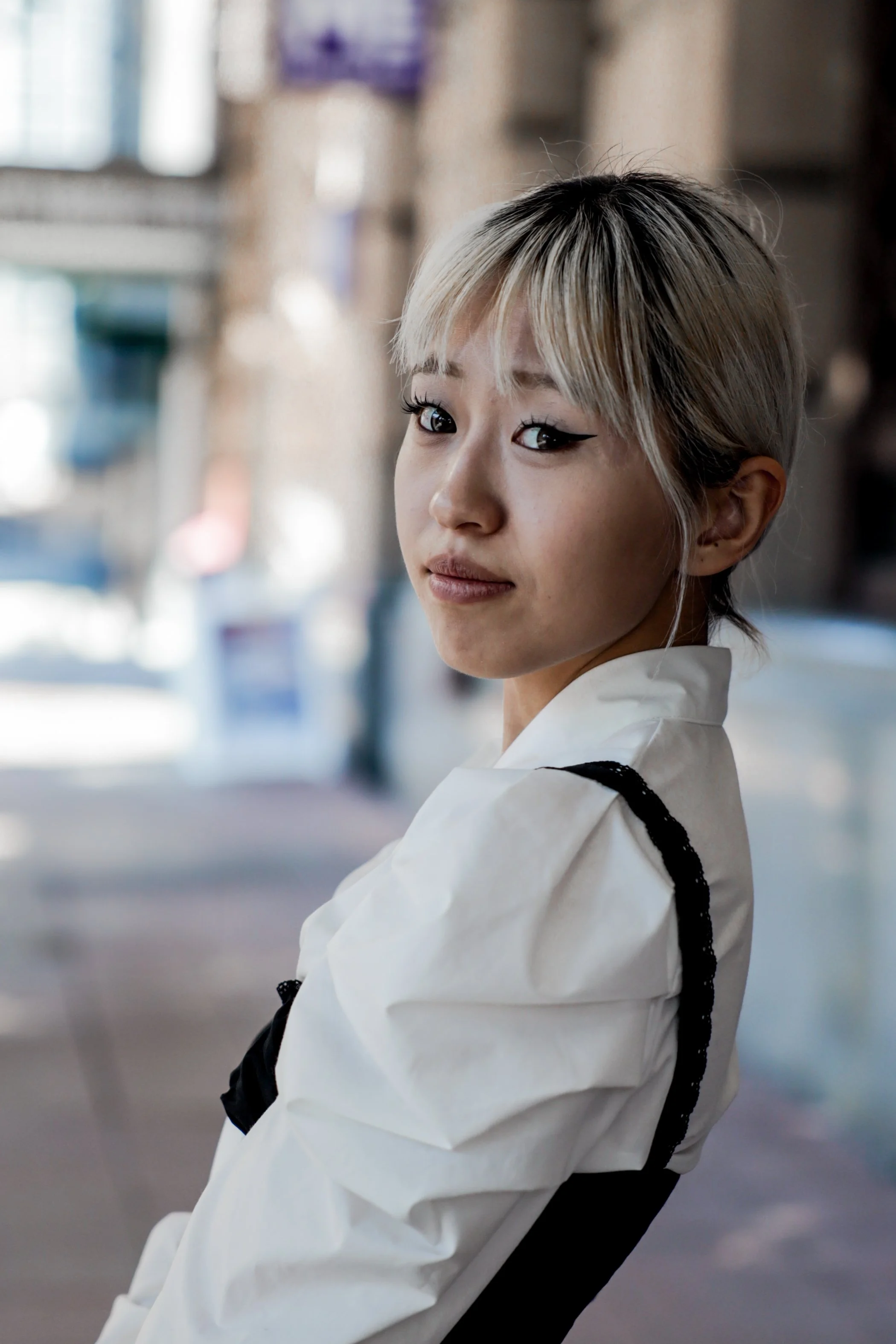 A young woman with short, blonde hair and winged eyeliner standing outdoors, looking over her shoulder at the camera, wearing a white and black jacket.