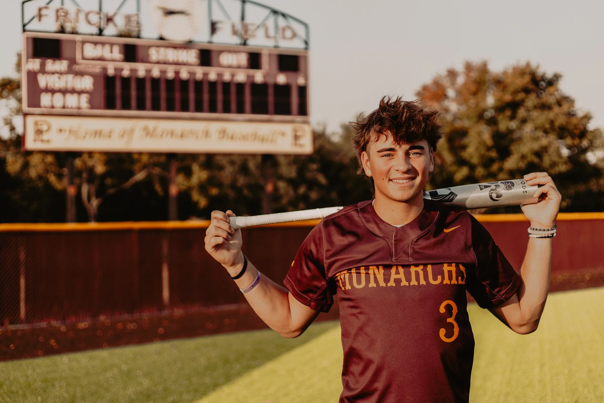 A young male athlete with curly brown hair smiling while holding a baseball bat over his shoulder, standing on a baseball field with an outfield fence and a scoreboard in the background during sunset.