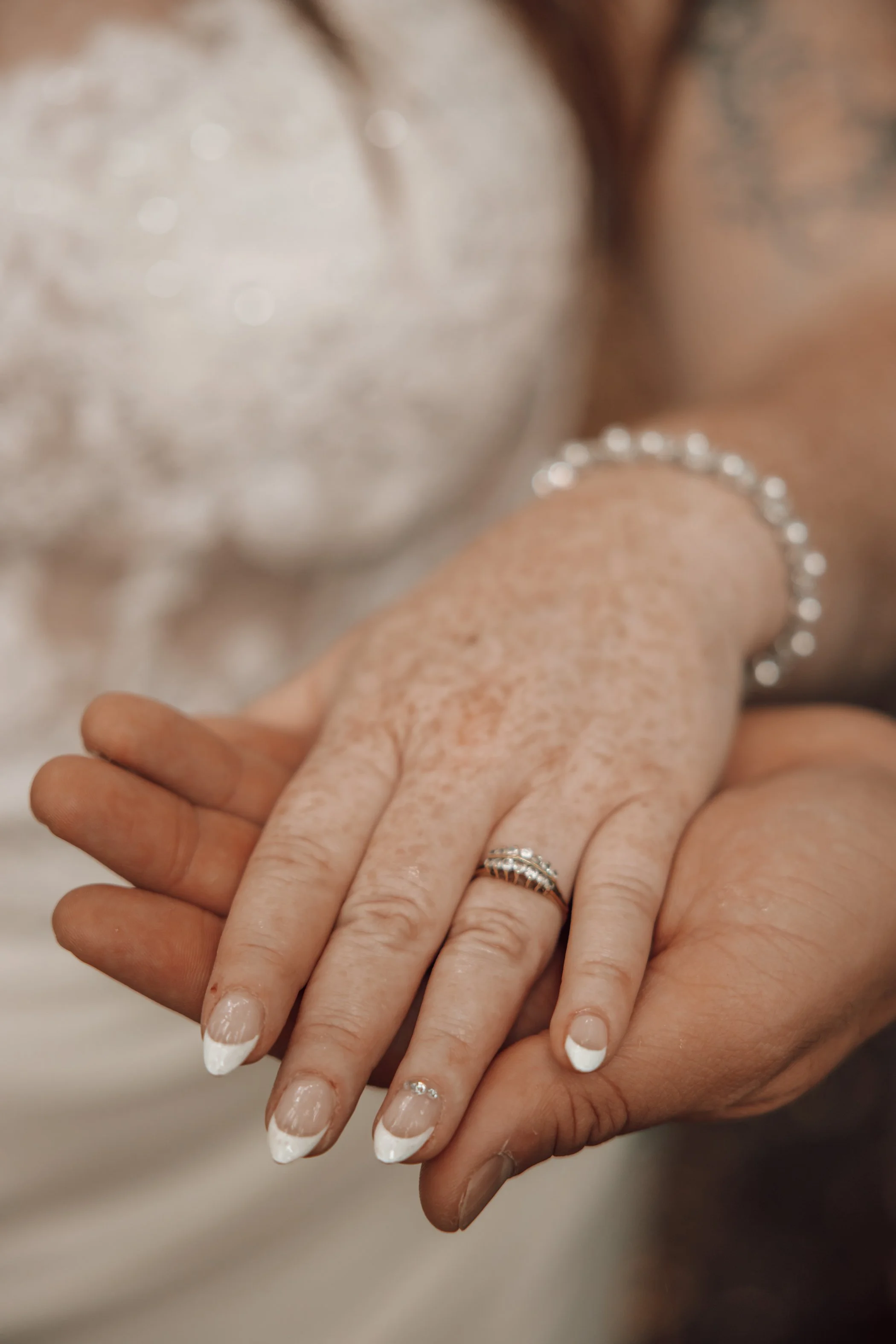 Close-up of two hands, one adult and one elderly, with the elderly hand resting on top of the adult hand. The adult has manicured nails with French tips and jewelry on the fingers and wrist.