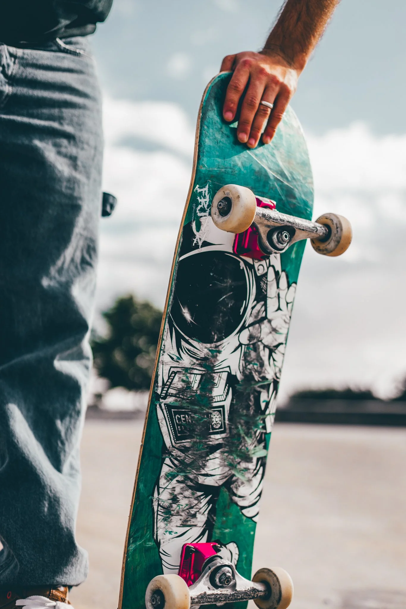 Close-up of a person holding a skateboard with a graphic design, wearing gray pants, with the background showing a cloudy sky and an open outdoor area.