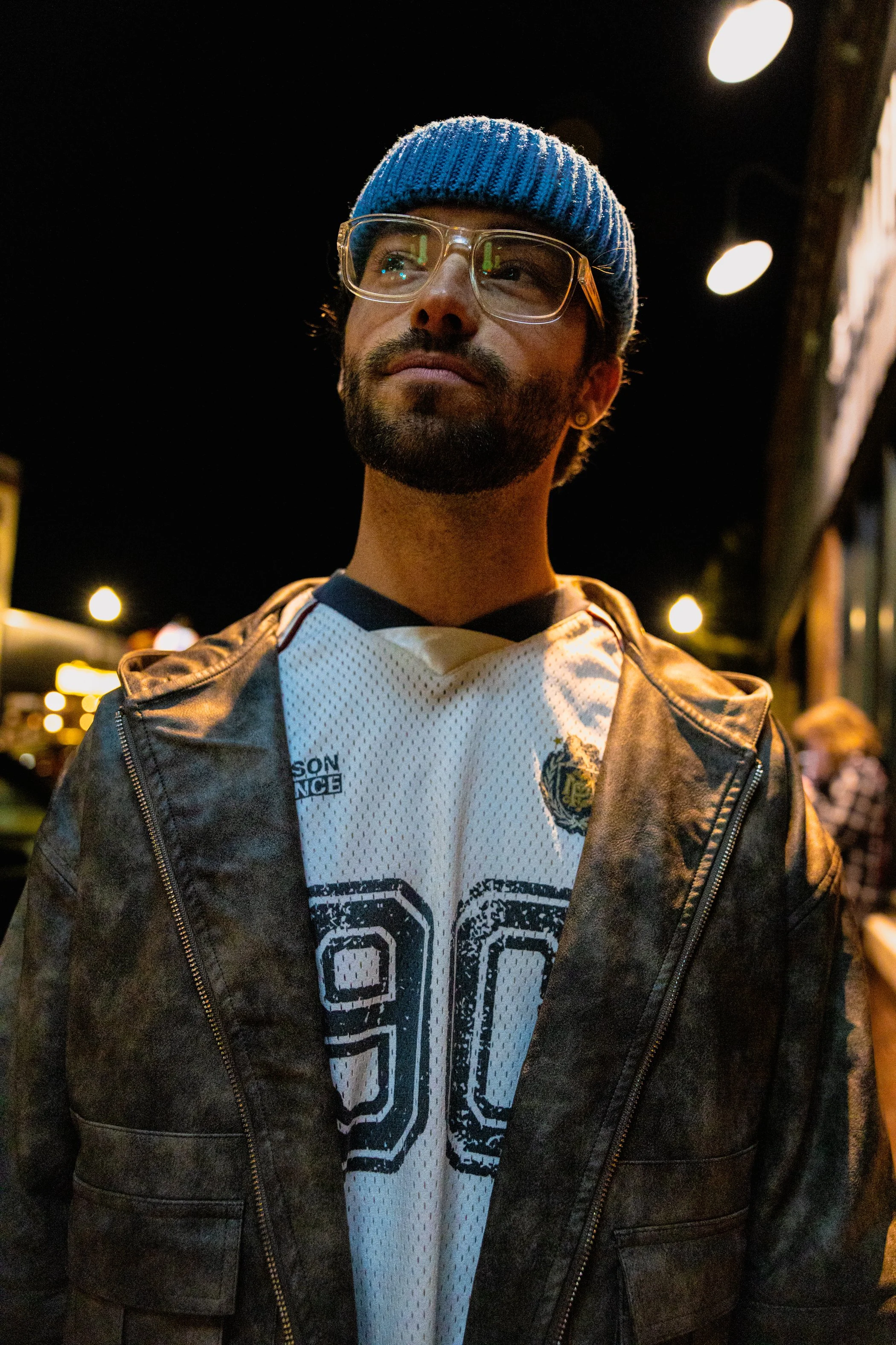 A man with glasses, a blue knit hat, and a beard standing outdoors at night with blurred city lights in the background.