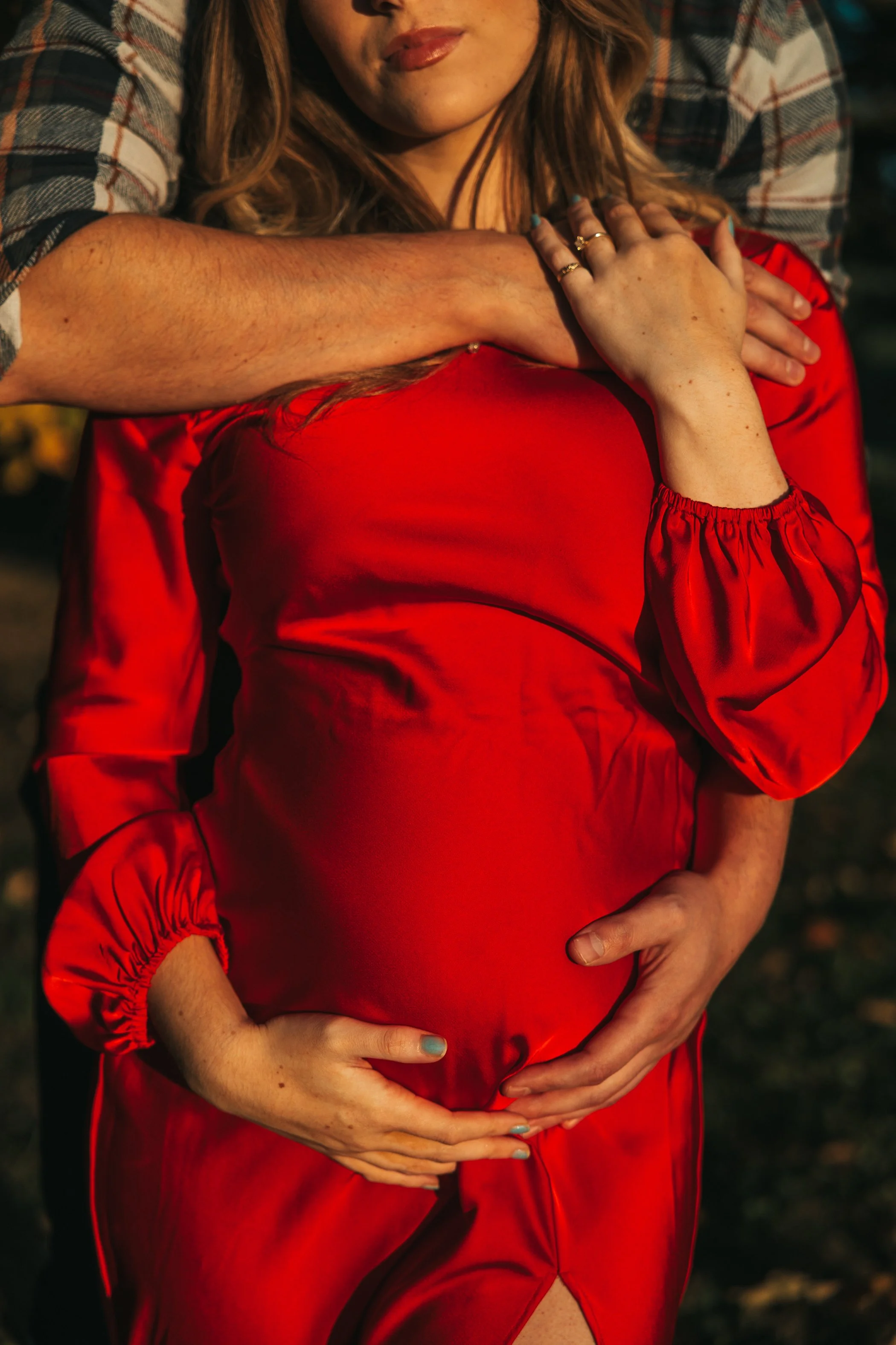 A pregnant woman wearing a red satin dress and gold rings, holding her belly with both hands, and an older person with a plaid shirt wrapping their arm around her shoulders, also placing a hand on her belly.