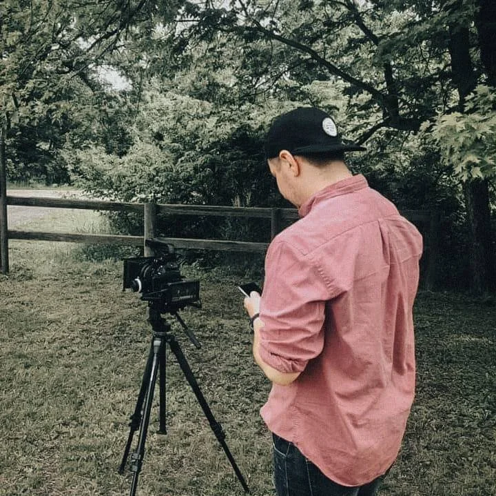 Person wearing a pink shirt and black cap standing outdoors, using a smartphone near a video camera on a tripod, with trees and a wooden fence in the background.