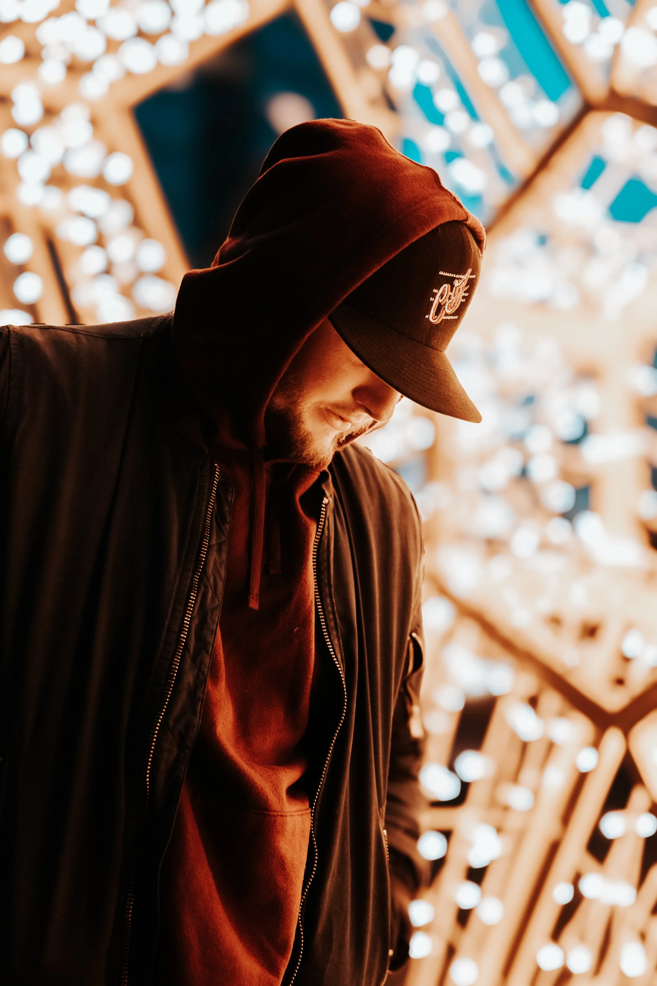 A young man is wearing a dark hoodie and baseball cap, looking down with soft lighting, in front of a blurred background with bright circular lights.