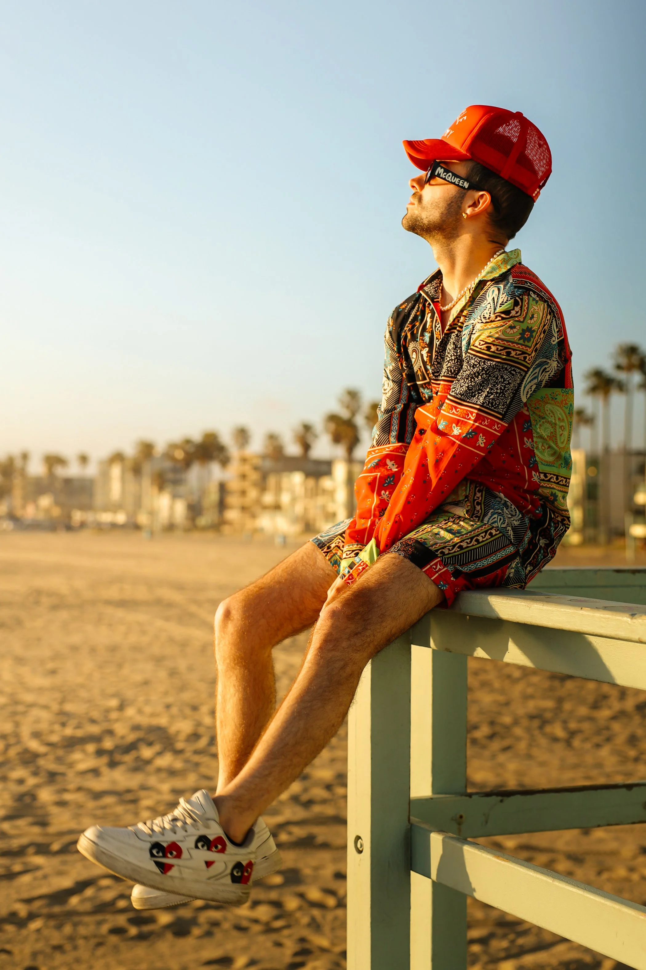 A man wearing a red and black patterned shirt, red baseball cap, black glasses with 'McQueen' written on them, and white sneakers with 'BAPE' logos, is sitting on a white railing at the beach with palm trees and buildings in the background during sun