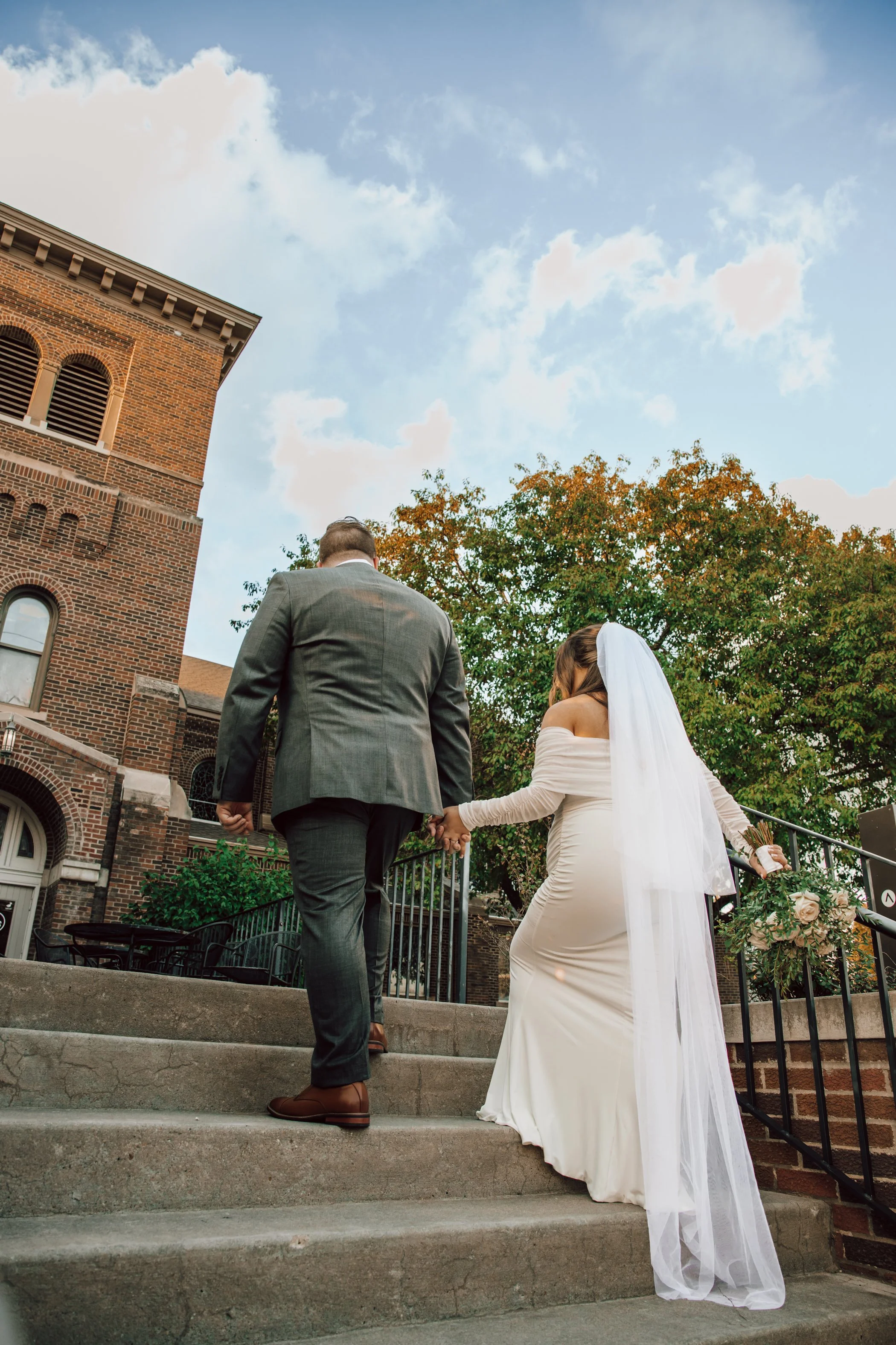 A couple getting married, dressed in wedding attire, walking up the steps of a brick building with a tower, holding hands, with a tree and clouds in the background.