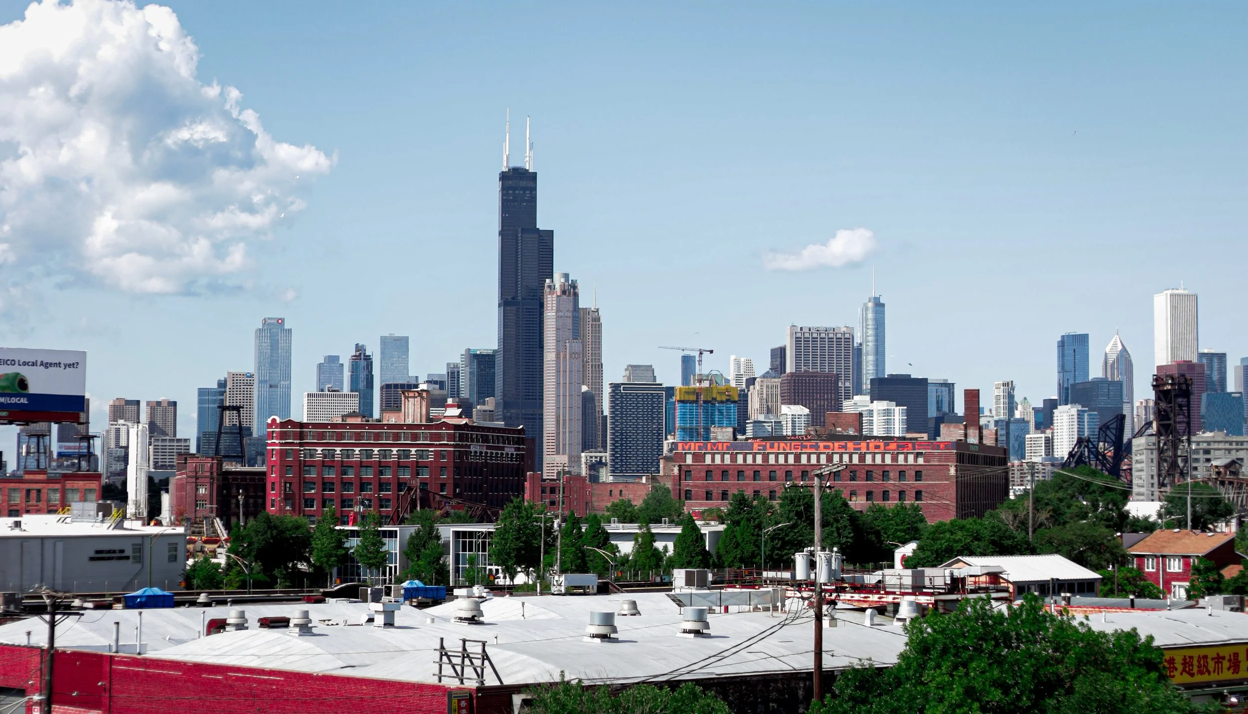 A city skyline featuring the Willis Tower with surrounding high-rise buildings under a partly cloudy sky.
