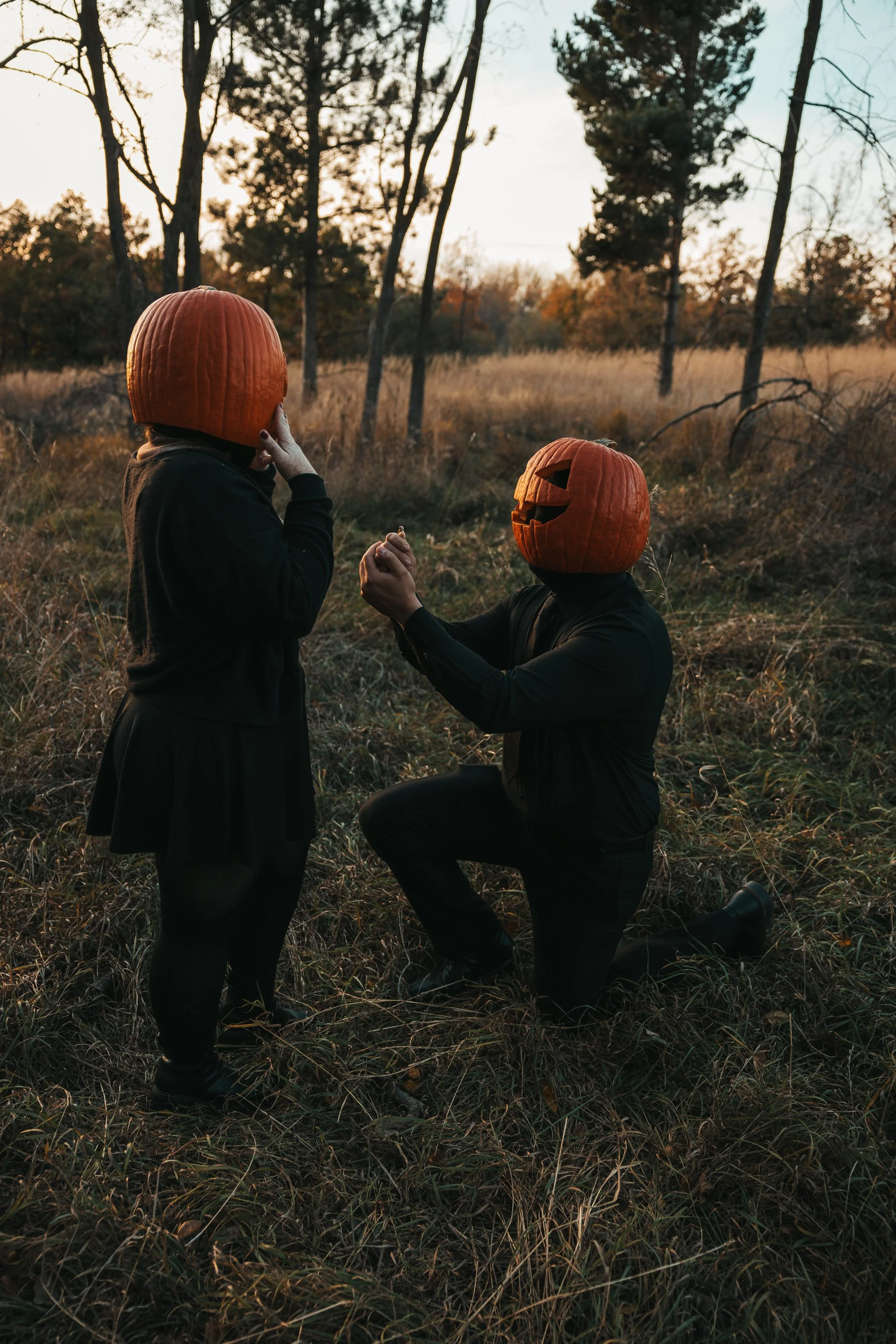 Two people in a wooded field during sunset, one kneeling and proposing with a pumpkin mask, the other standing with a pumpkin mask, holding hands.