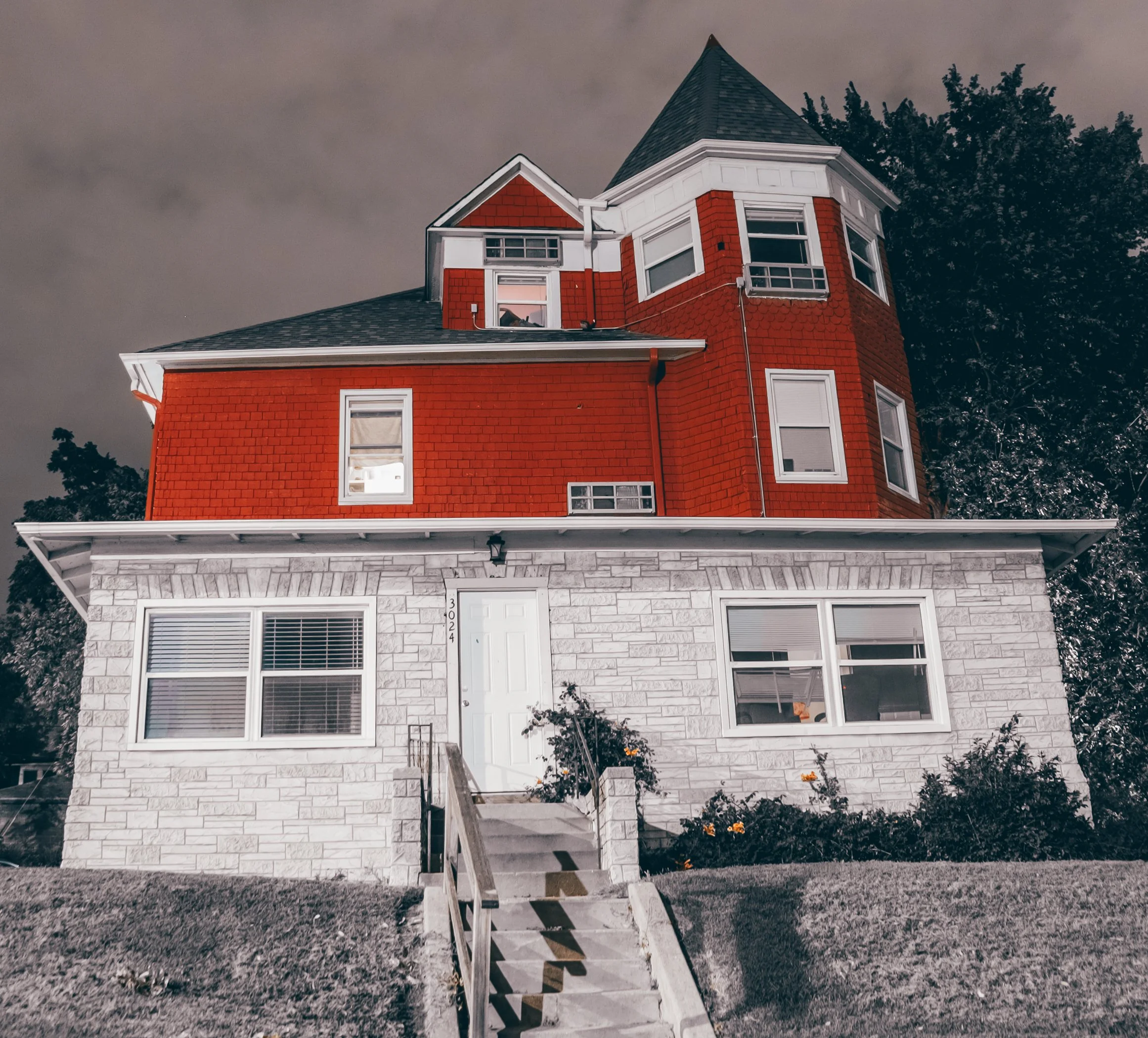 A two-story house with red brick siding on the upper level and white stone on the lower level, with multiple windows and a set of stairs leading to the front door, set against a cloudy sky with trees in the background.
