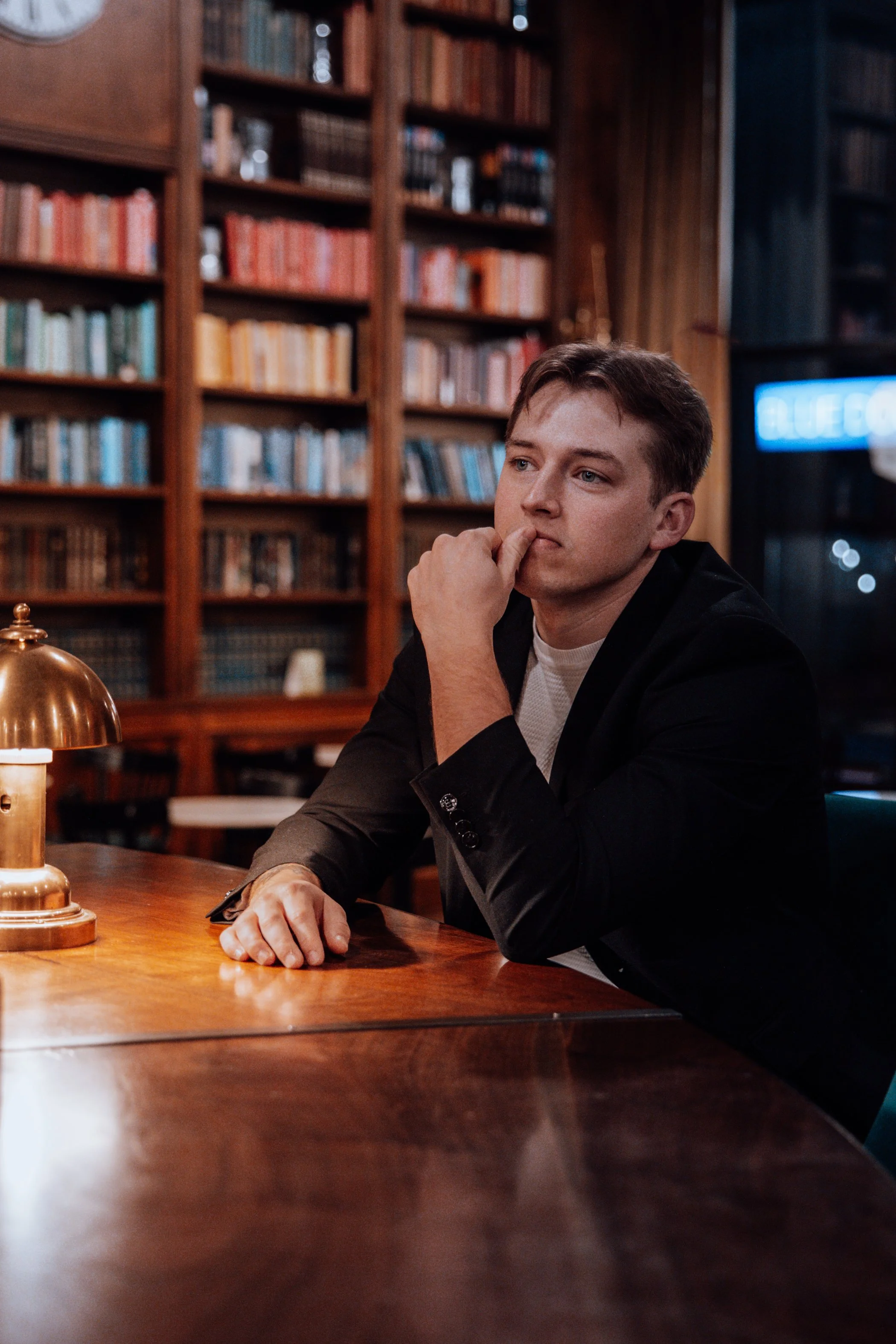 Young man sitting at a wooden table in a library or study room with filled bookshelves in the background, resting his chin on his hand, looking contemplative.