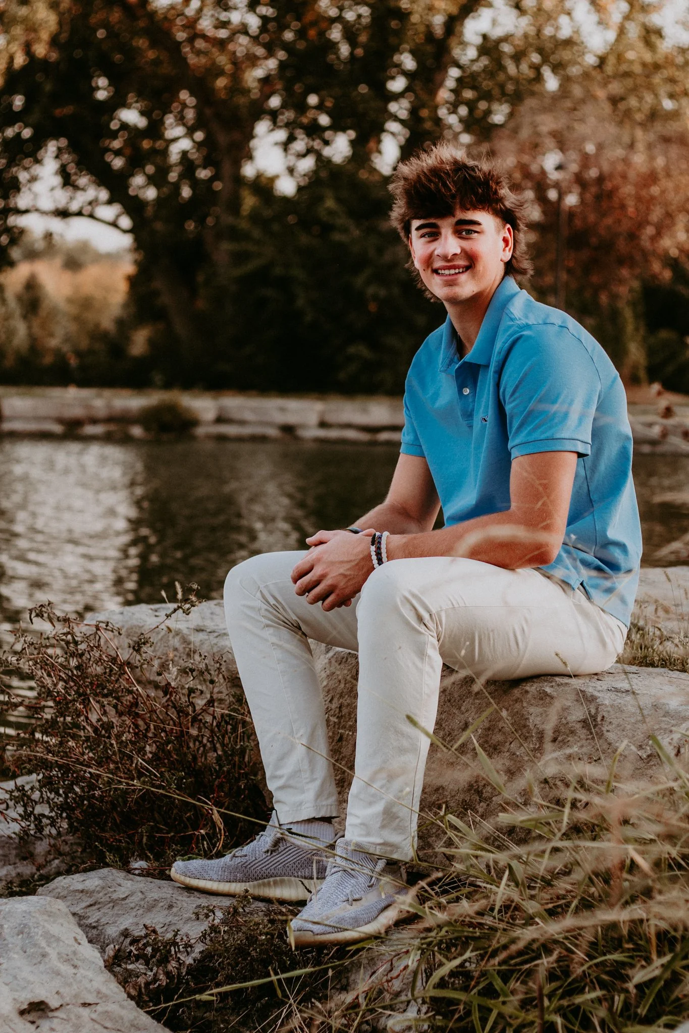 A young man with curly brown hair and a smile, wearing a blue polo shirt and white pants, sitting on rocks near a body of water with trees in the background during sunset.