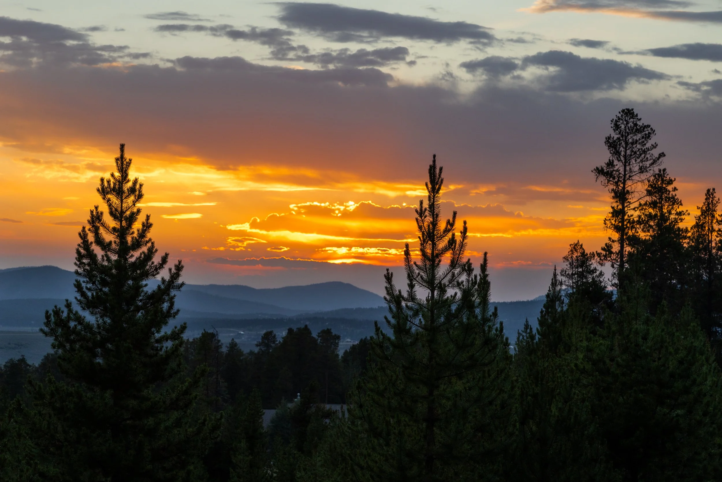 Sunset over a forested mountain landscape with pine trees in the foreground and layers of mountains in the background, dramatic sky with dark clouds and orange glow from the setting sun.