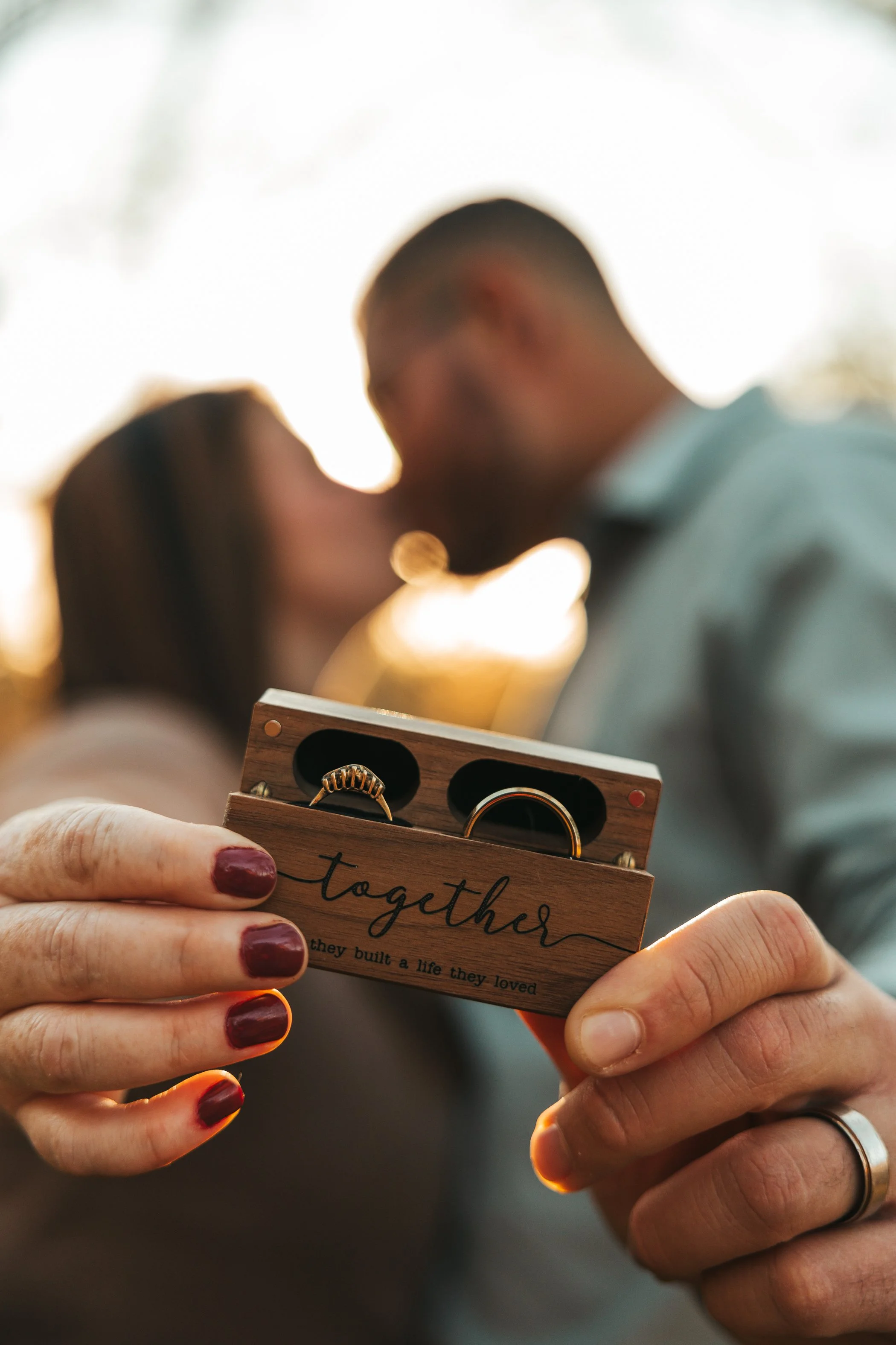 Close-up of a person's hands holding a small wooden box with rings inside. The box has the word 'together' written on it. In the background, a couple is slightly out of focus, sharing a kiss or close moment, with warm lighting.