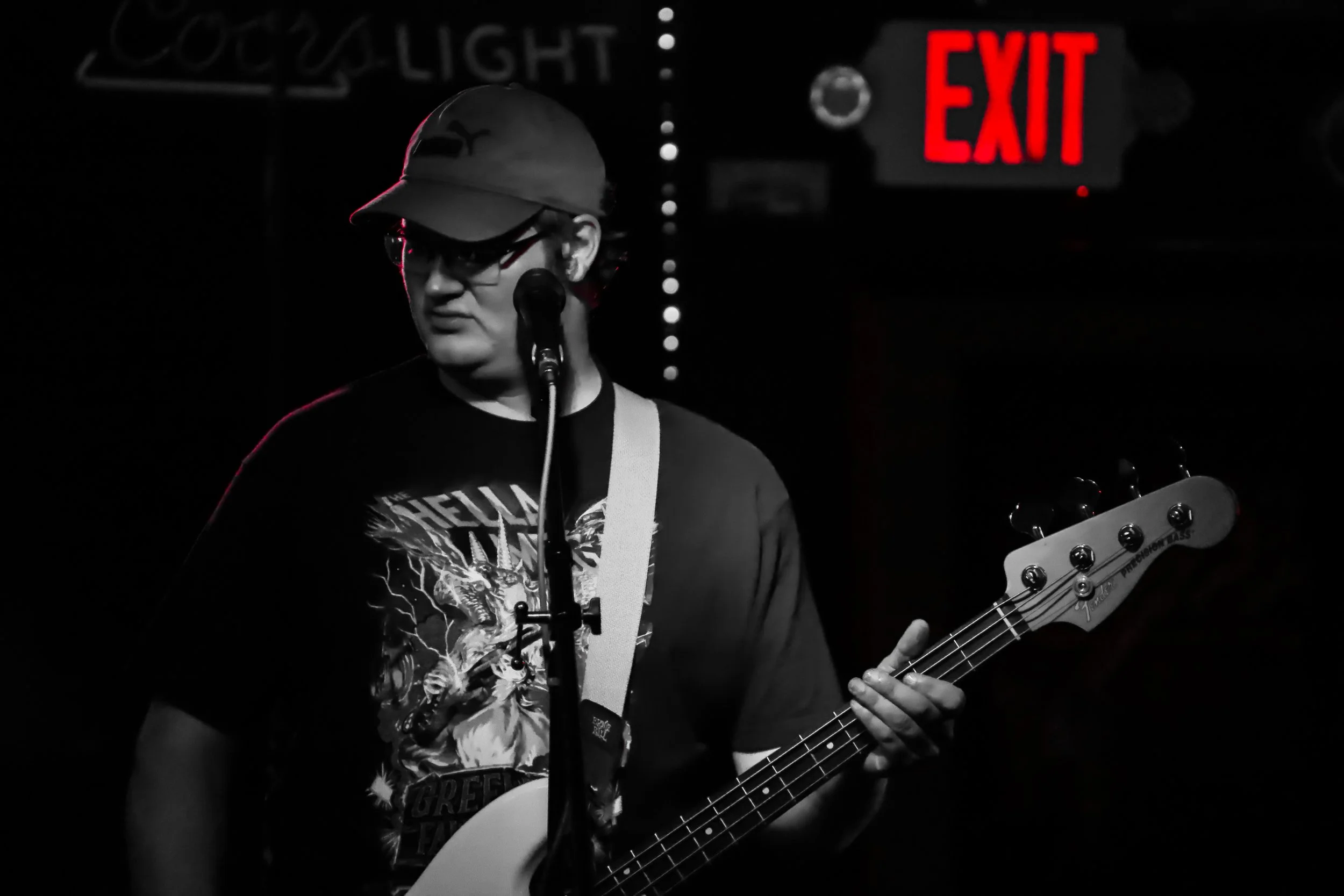 A person with glasses and a cap playing an electric guitar in a dark room with a bright red 'EXIT' sign and a partially visible drawing or word on the wall.