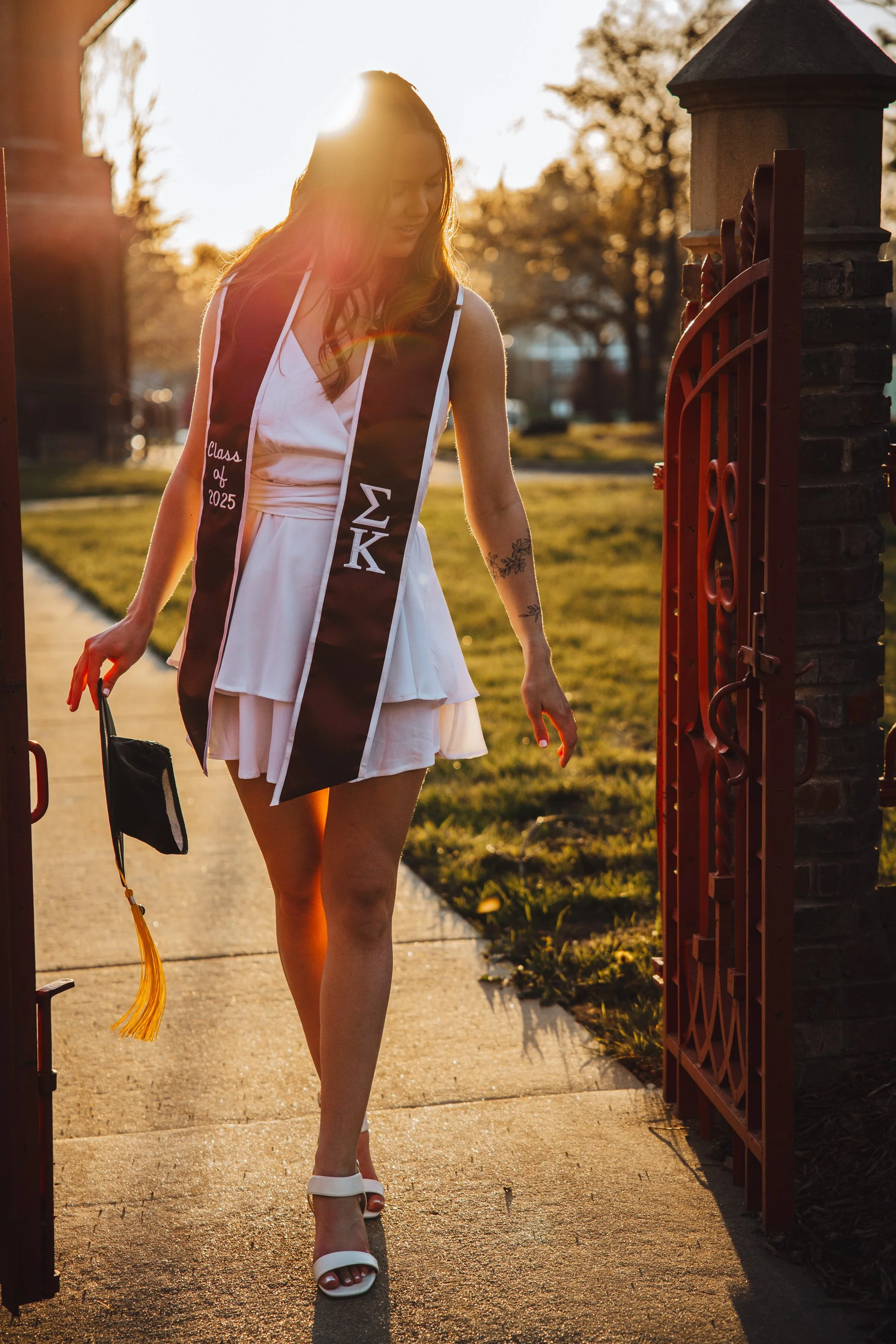 A woman wearing a white dress and high heels walking through a gate with the sunlight shining behind her during sunset.
