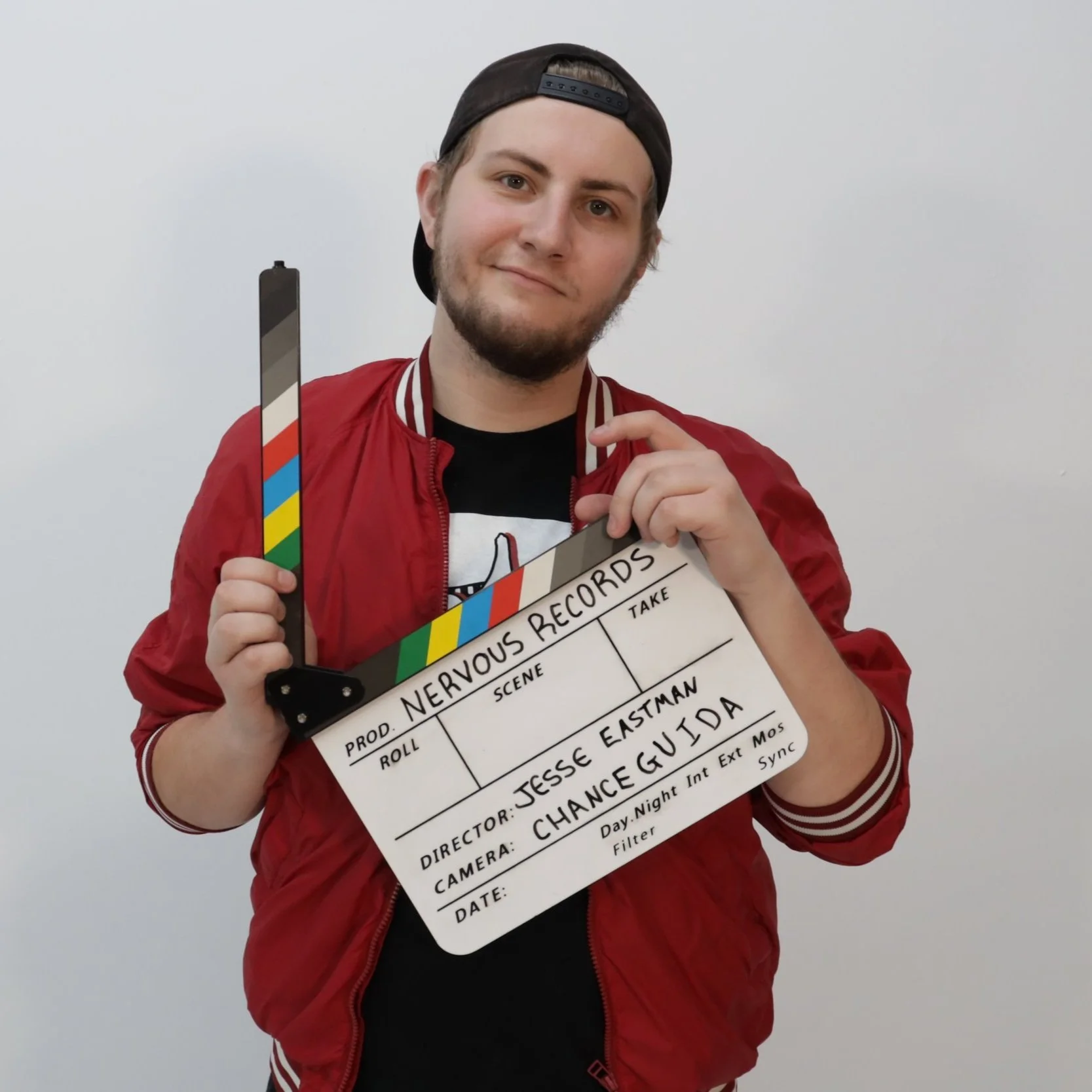 A young man wearing a black cap backward and a red jacket holds a clapperboard. The background is plain white.