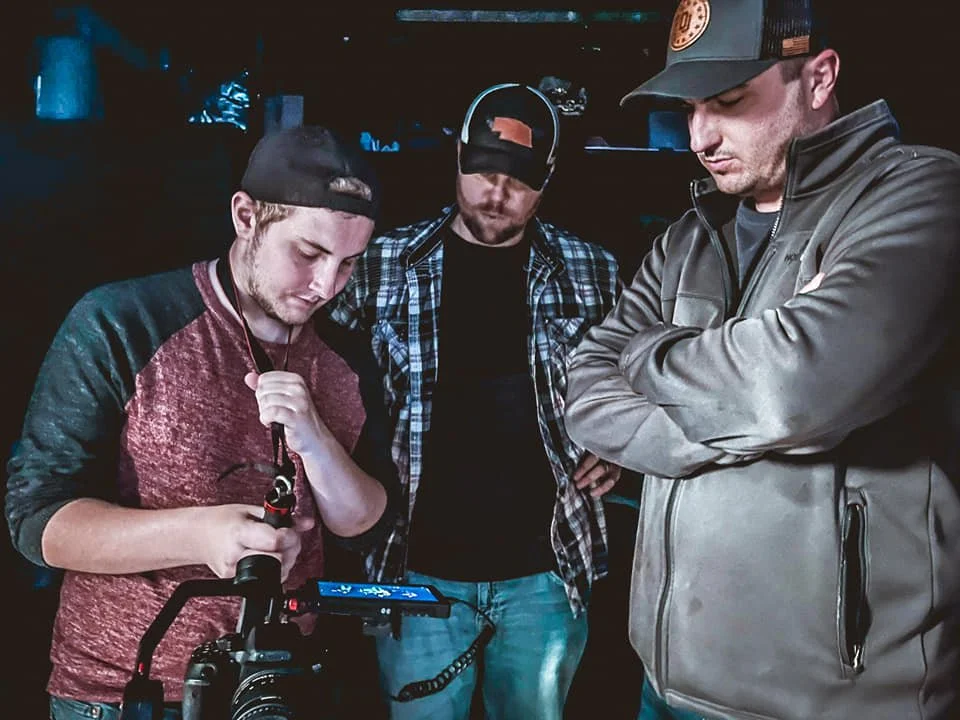 Three men are gathered around a camera, with one of them adjusting the settings while the other two observe attentively, in a dimly lit setting.