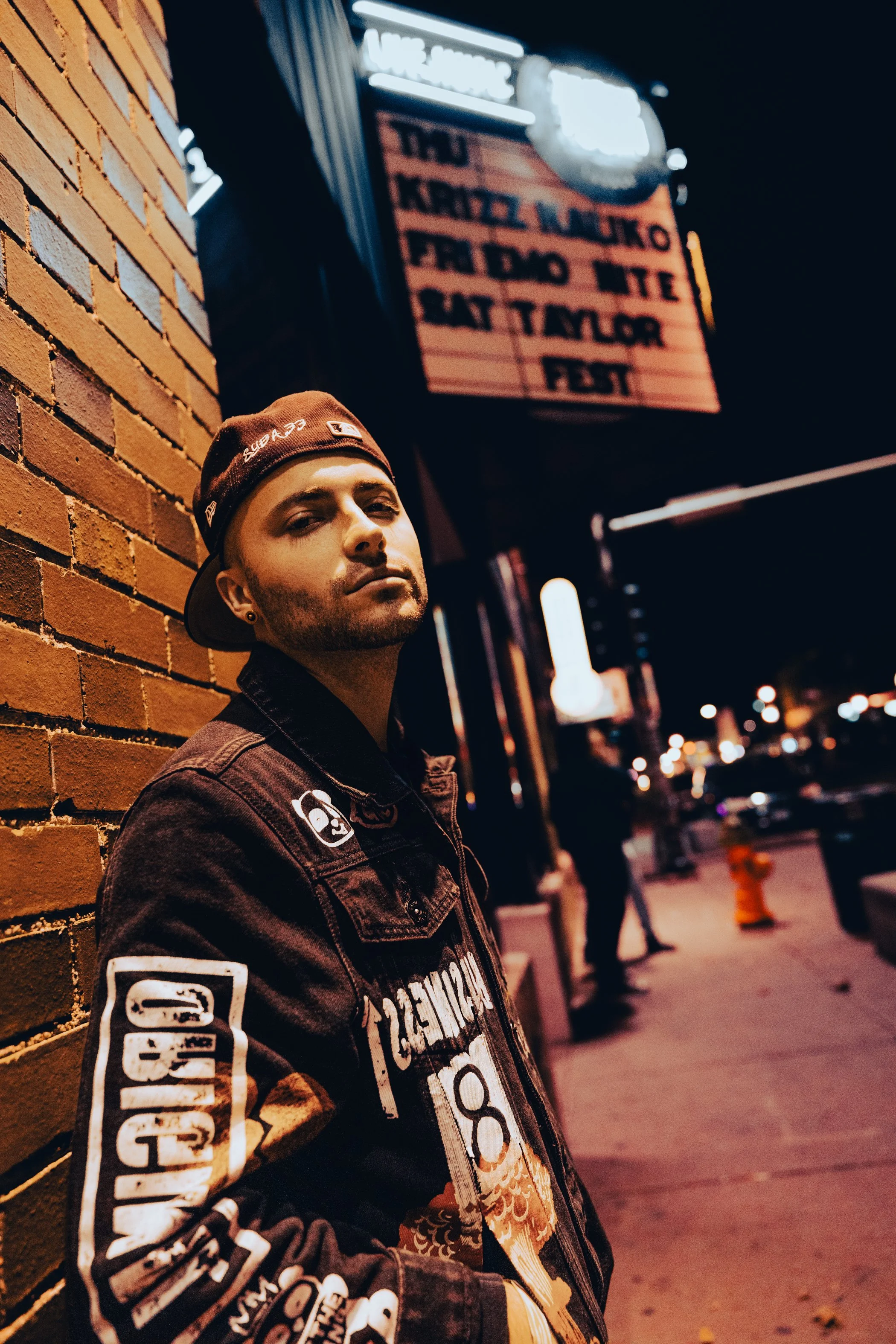 A man with dark hair and a goatee wearing a black baseball cap worn backwards, a black denim jacket with various patches, standing against a brick wall on a city street at night, with a neon sign and illuminated storefronts in the background.
