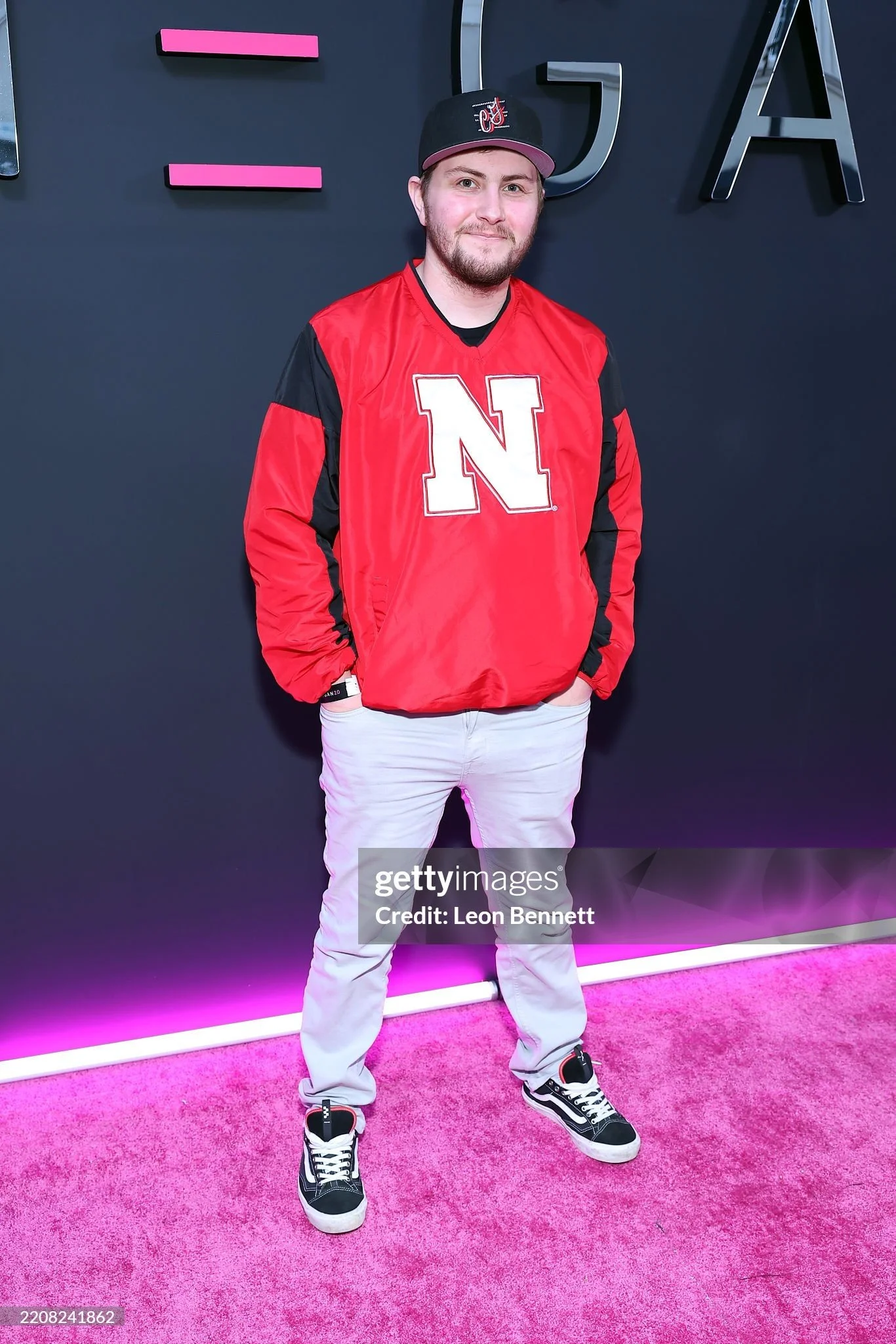 A man dressed in a red and black Nebraska Cornhuskers jacket, white pants, and black sneakers standing on a pink carpet in front of a dark wall with large metallic letters and pink accents.