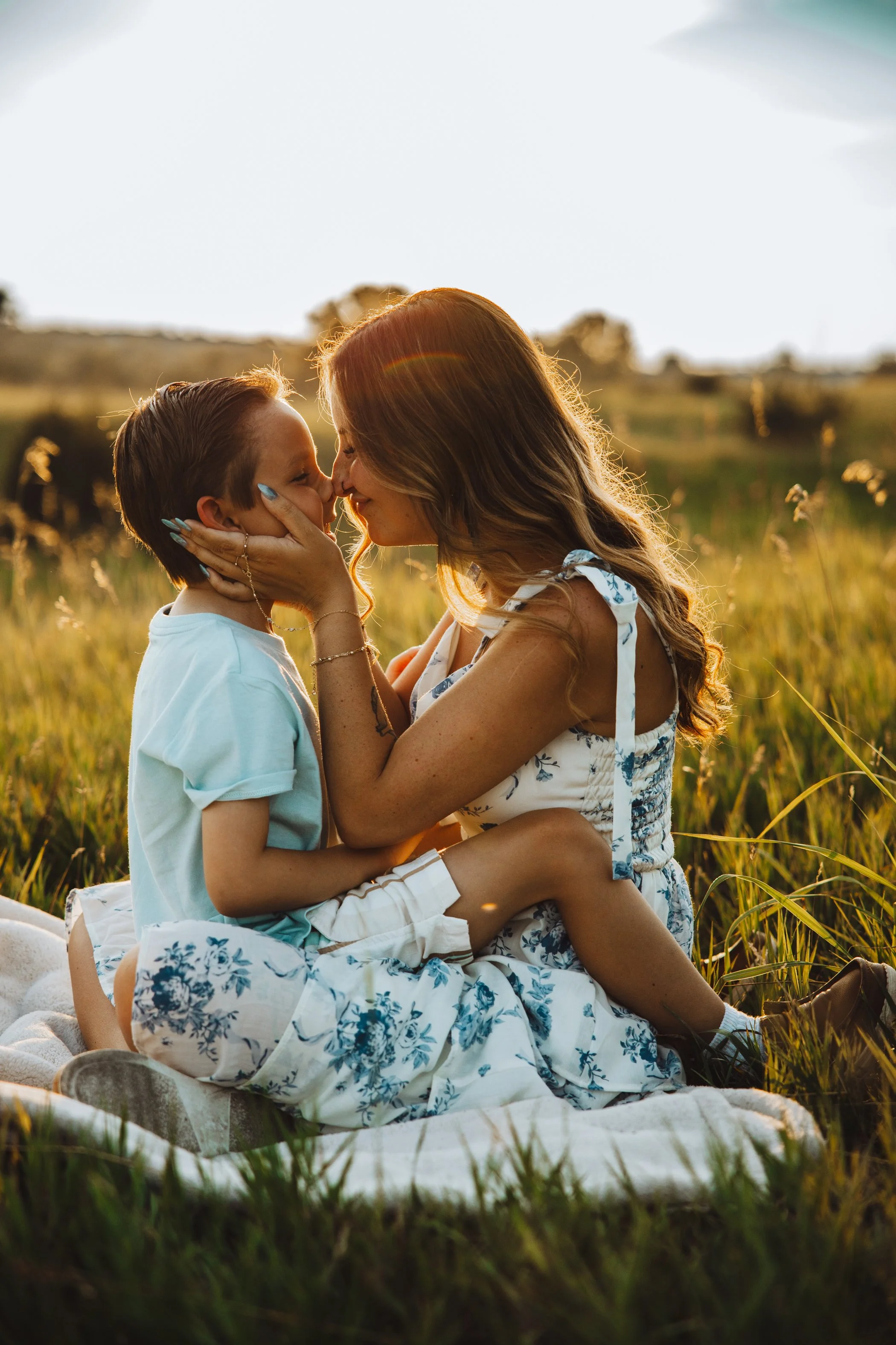 A woman and a boy share a tender moment, touching noses and smiling in a grassy field at sunset, with warm sunlight and a blurred background.