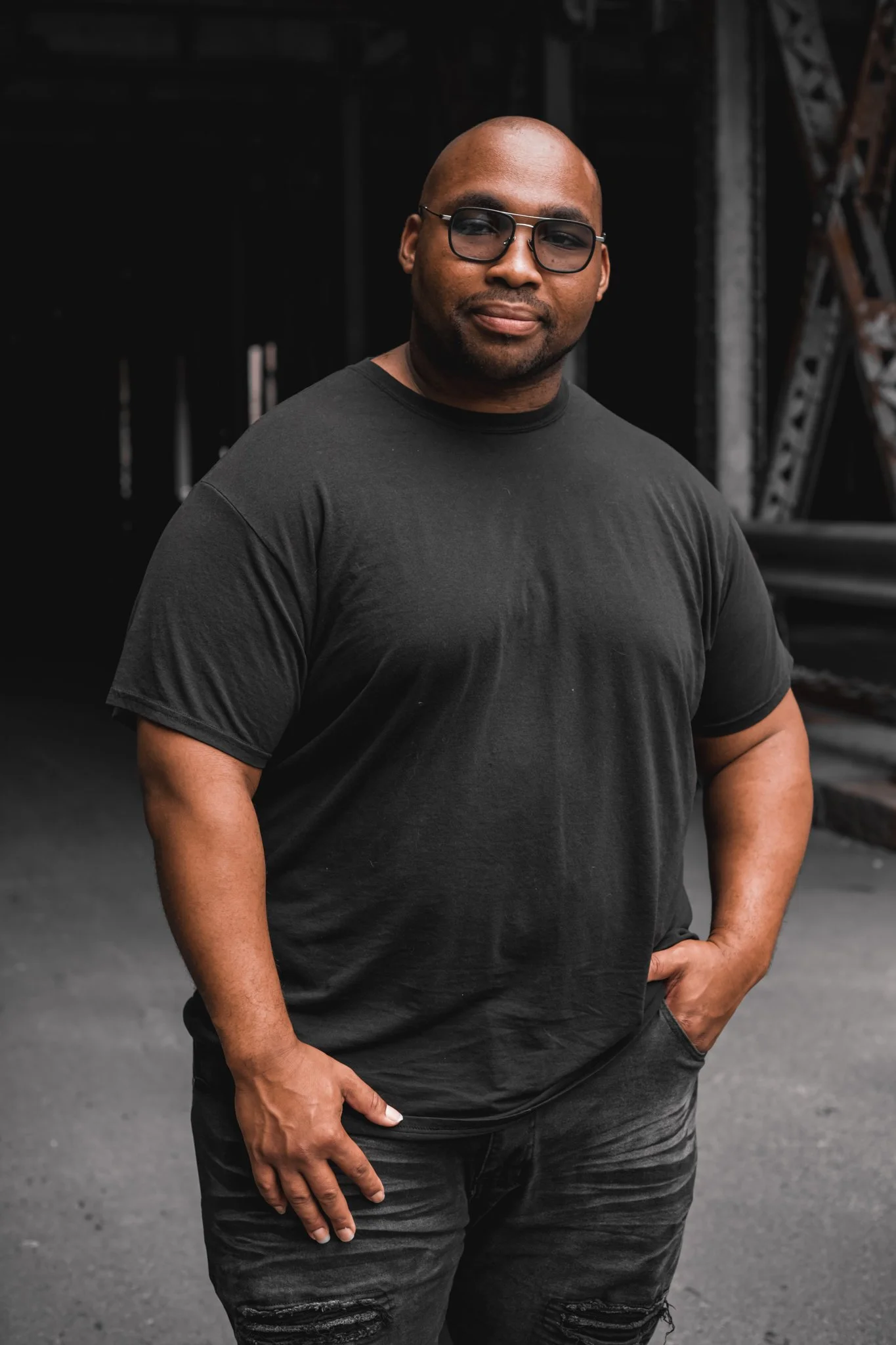 A man with dark skin, bald head, wearing black glasses, black t-shirt, and black ripped jeans standing outdoors in front of a dark background.