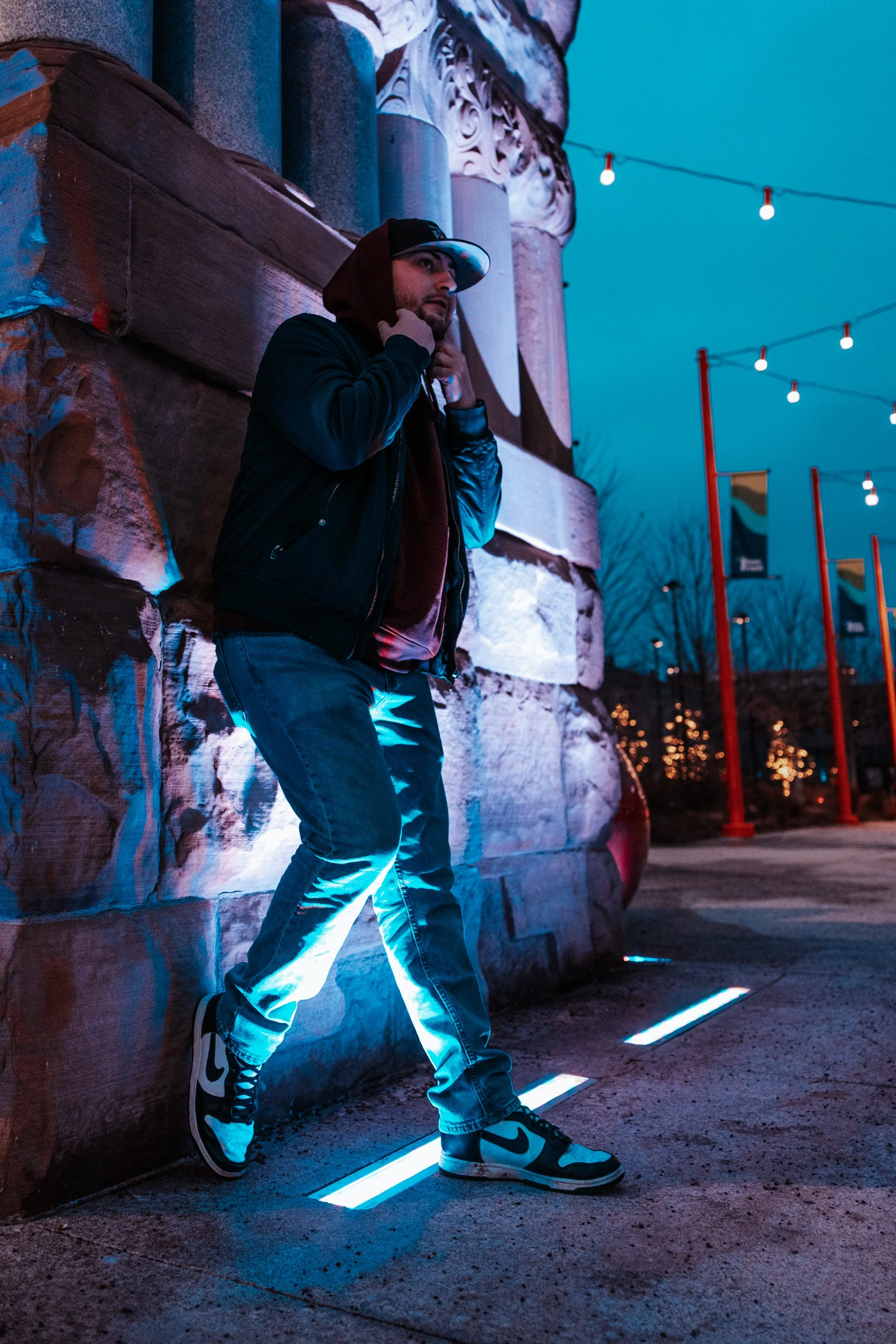 A man standing against a stone wall at night, illuminated by blue and purple lighting, wearing a black jacket, blue jeans, and Nike sneakers, with string lights overhead.