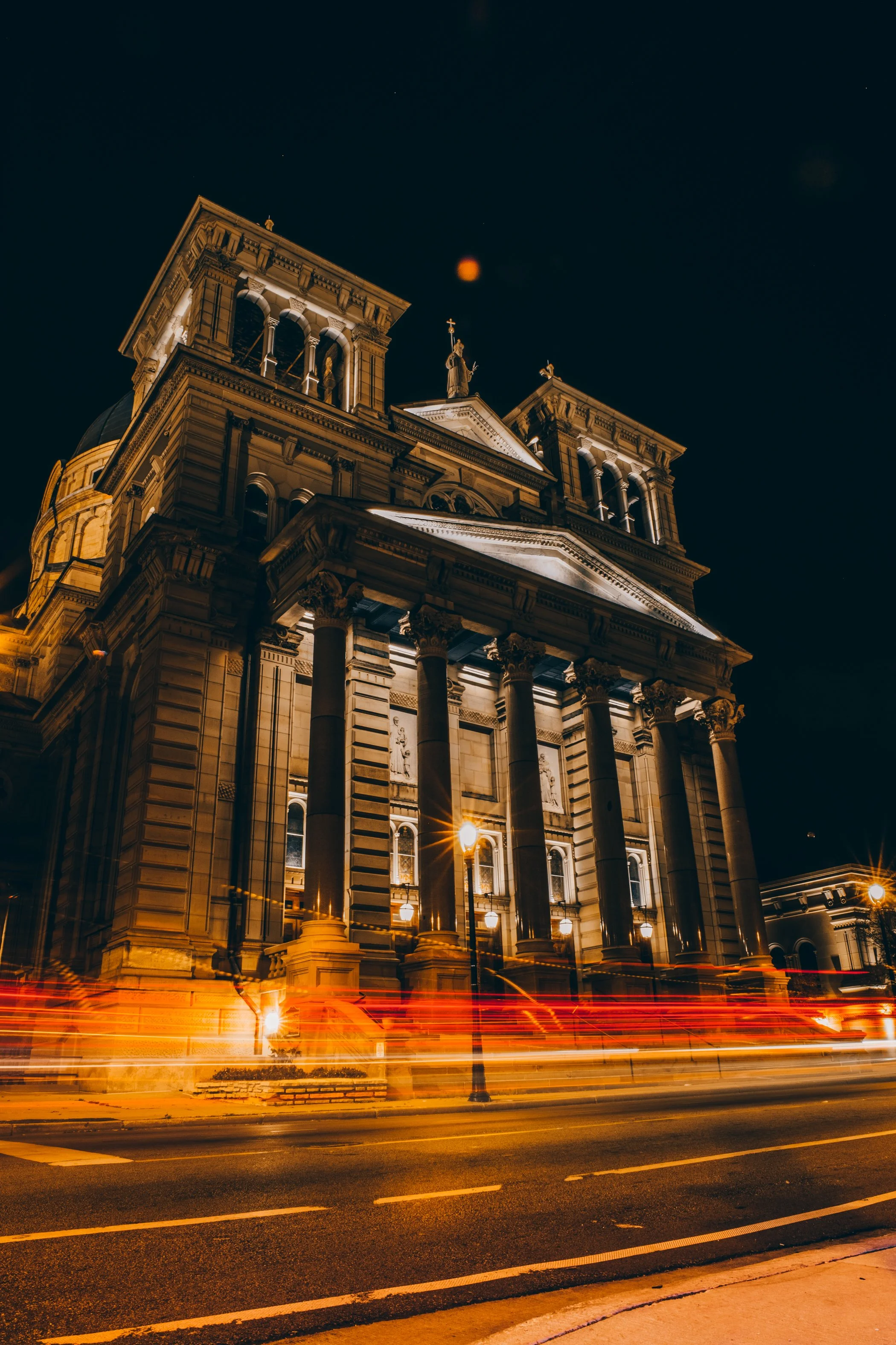 A historic building illuminated at night with classical architectural details and statues on top; light trails from passing vehicles in the street in front.