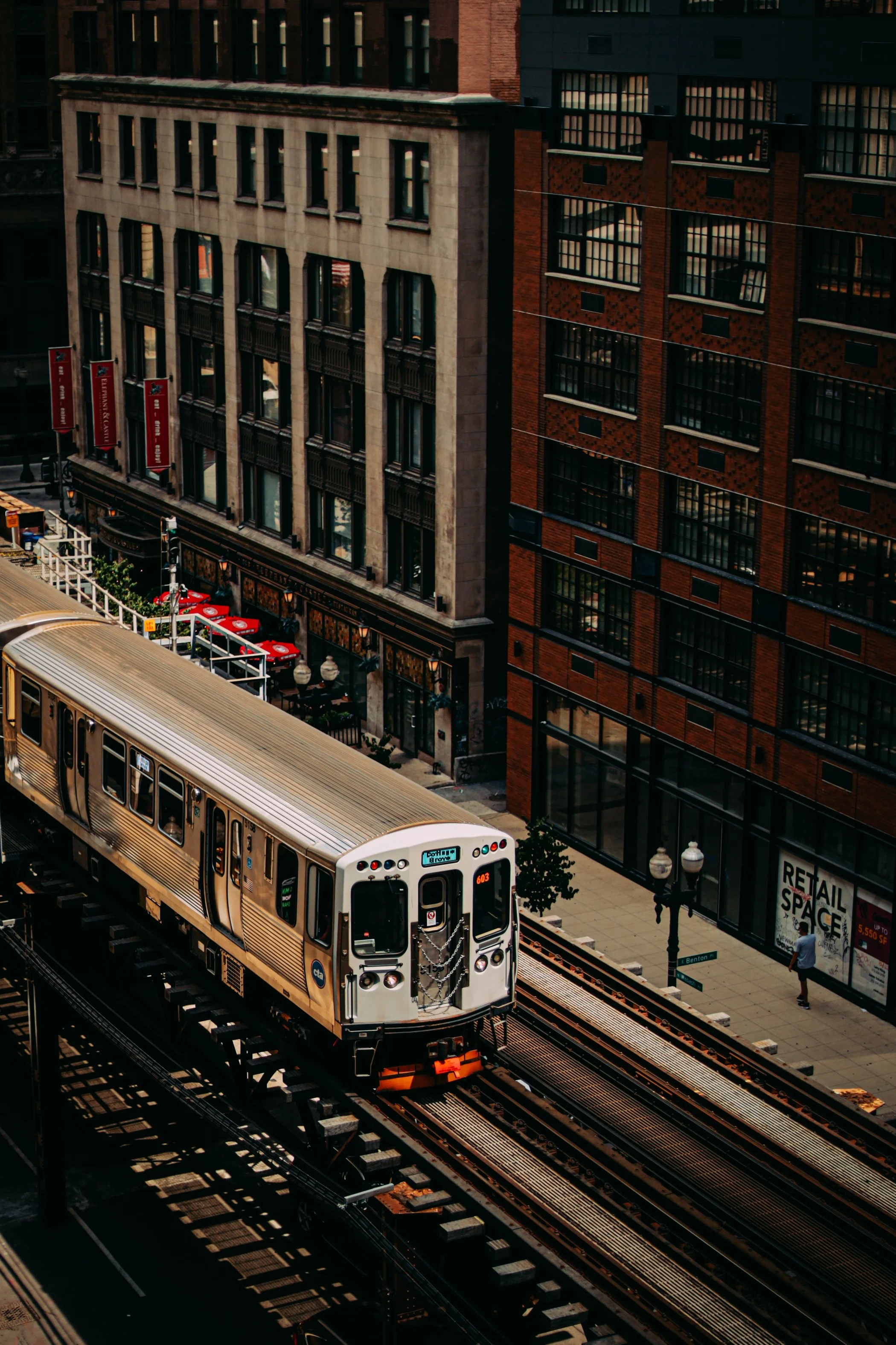 Urban scene with a yellow train on elevated tracks passing through city buildings, including brick and stone structures, with street pedestrians and storefronts visible.