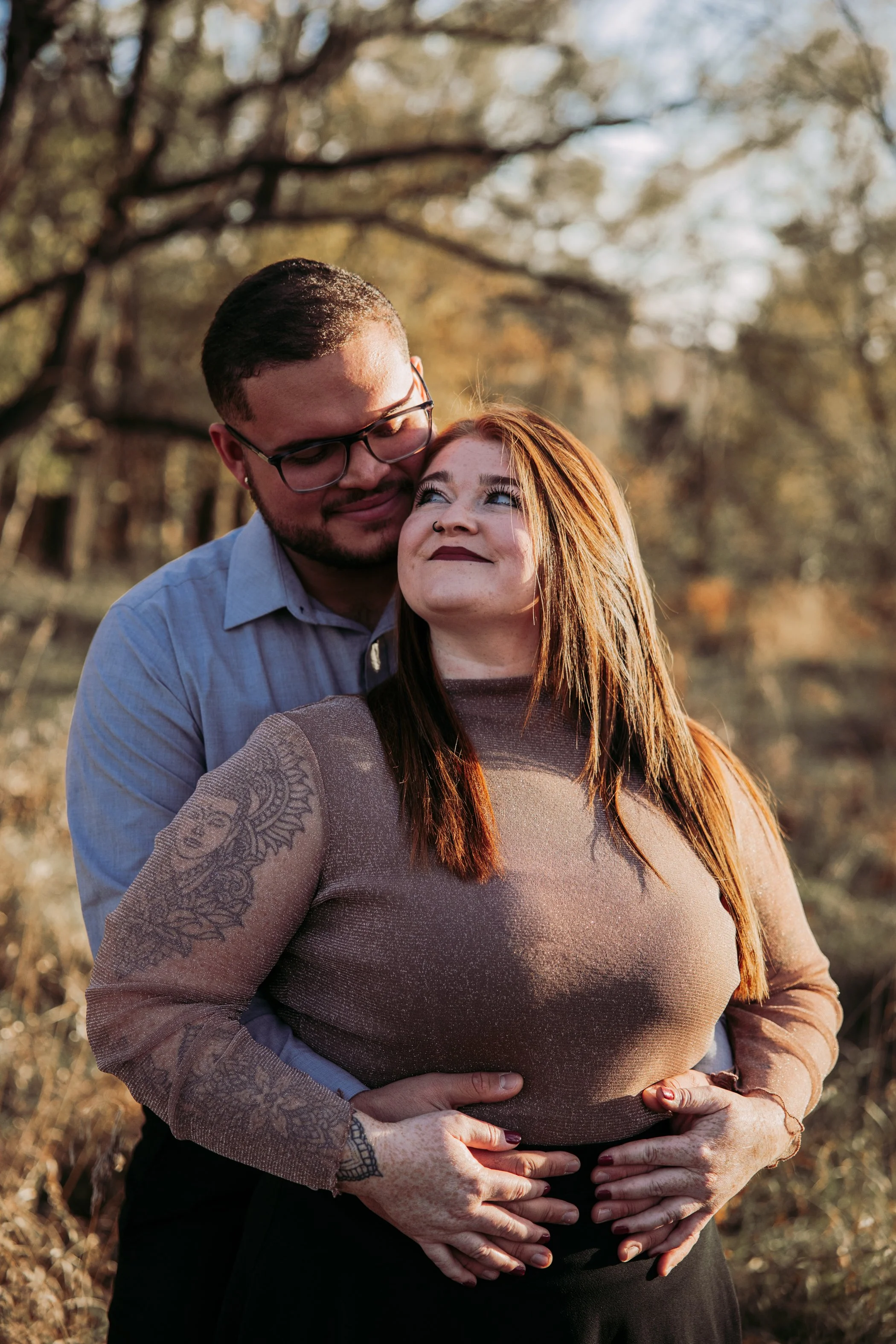 A man and woman stand close together outdoors in a wooded area with autumn foliage. The man is cuddling the woman from behind, both smiling and looking at each other affectionately. The woman has reddish-brown hair, a tattoo on her left arm, and is w