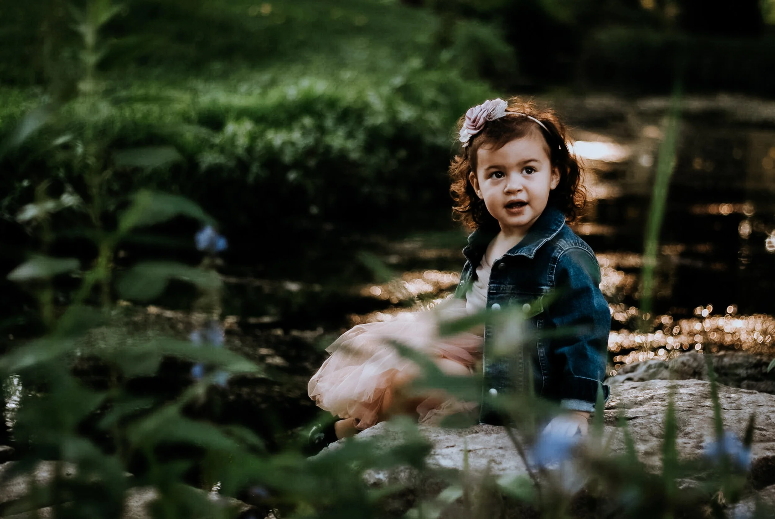 A young girl with curly brown hair and a pink flower headband sitting outdoors among green foliage, wearing a denim jacket and a light pink dress, looking surprised or curious.