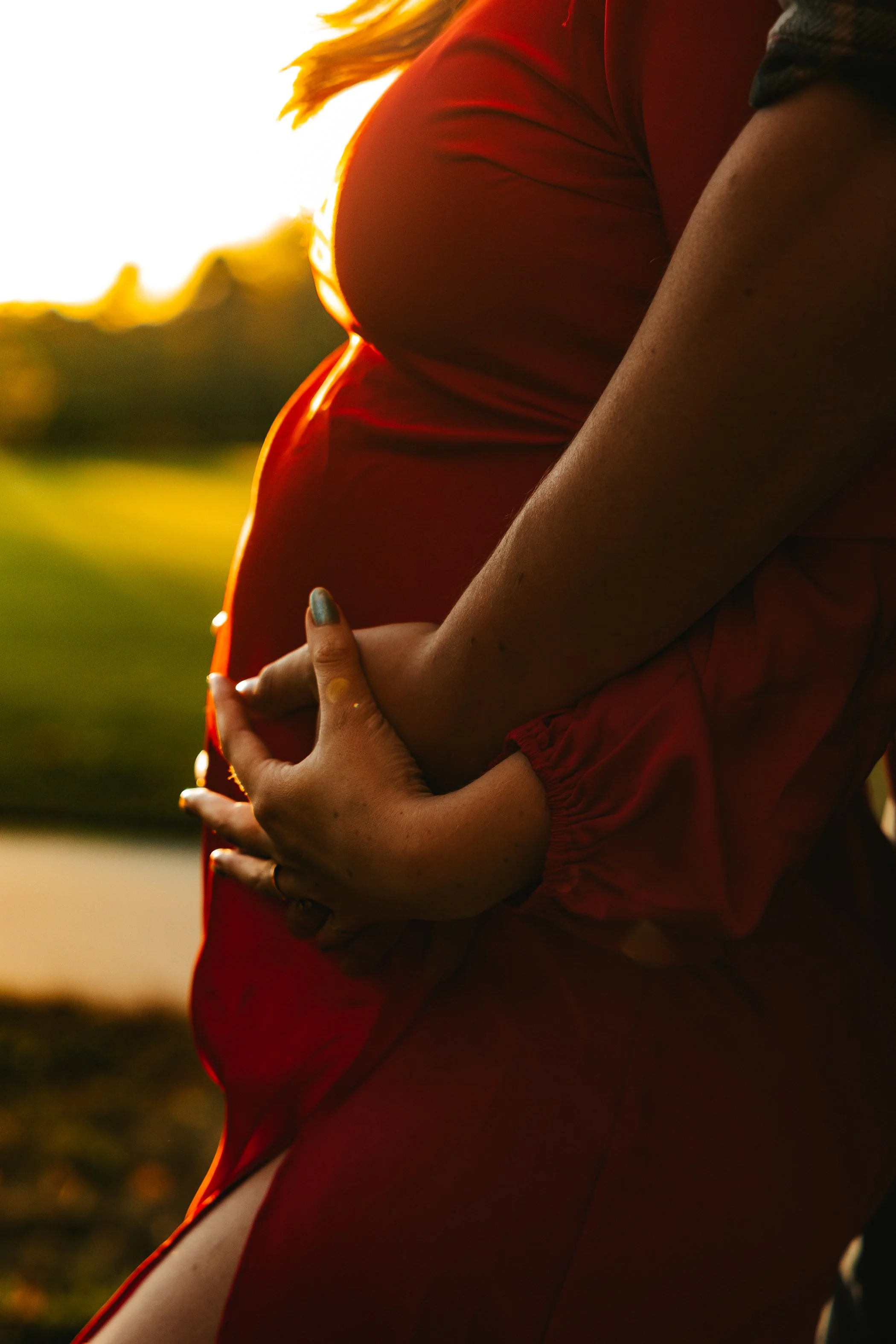 A pregnant person embracing their belly outdoors during sunset, wearing a red top, with the sun setting behind them.