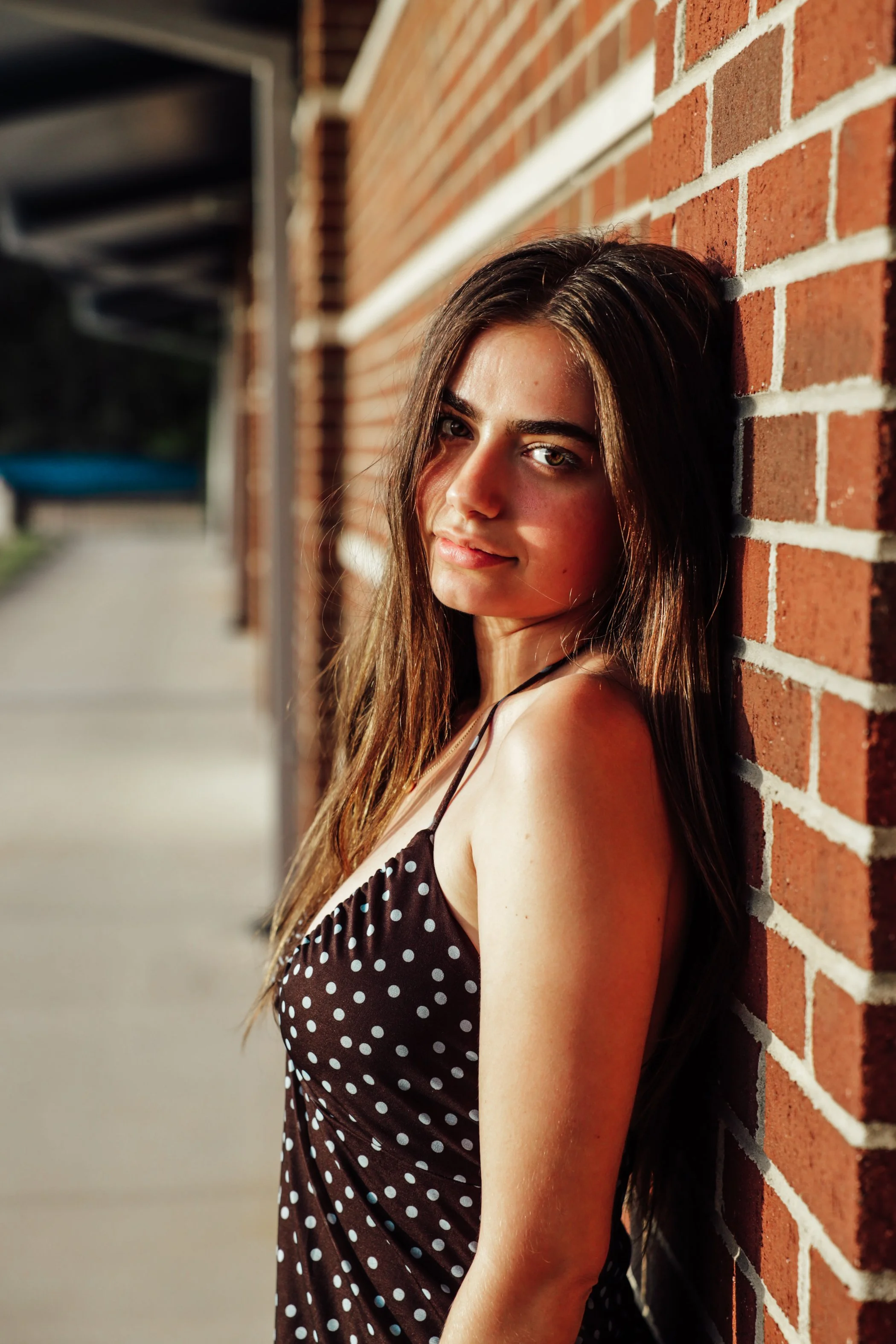 A young woman with long brown hair and light eyes leaning against a brick wall in sunlight, wearing a black dress with white polka dots and a black strap on her shoulder.